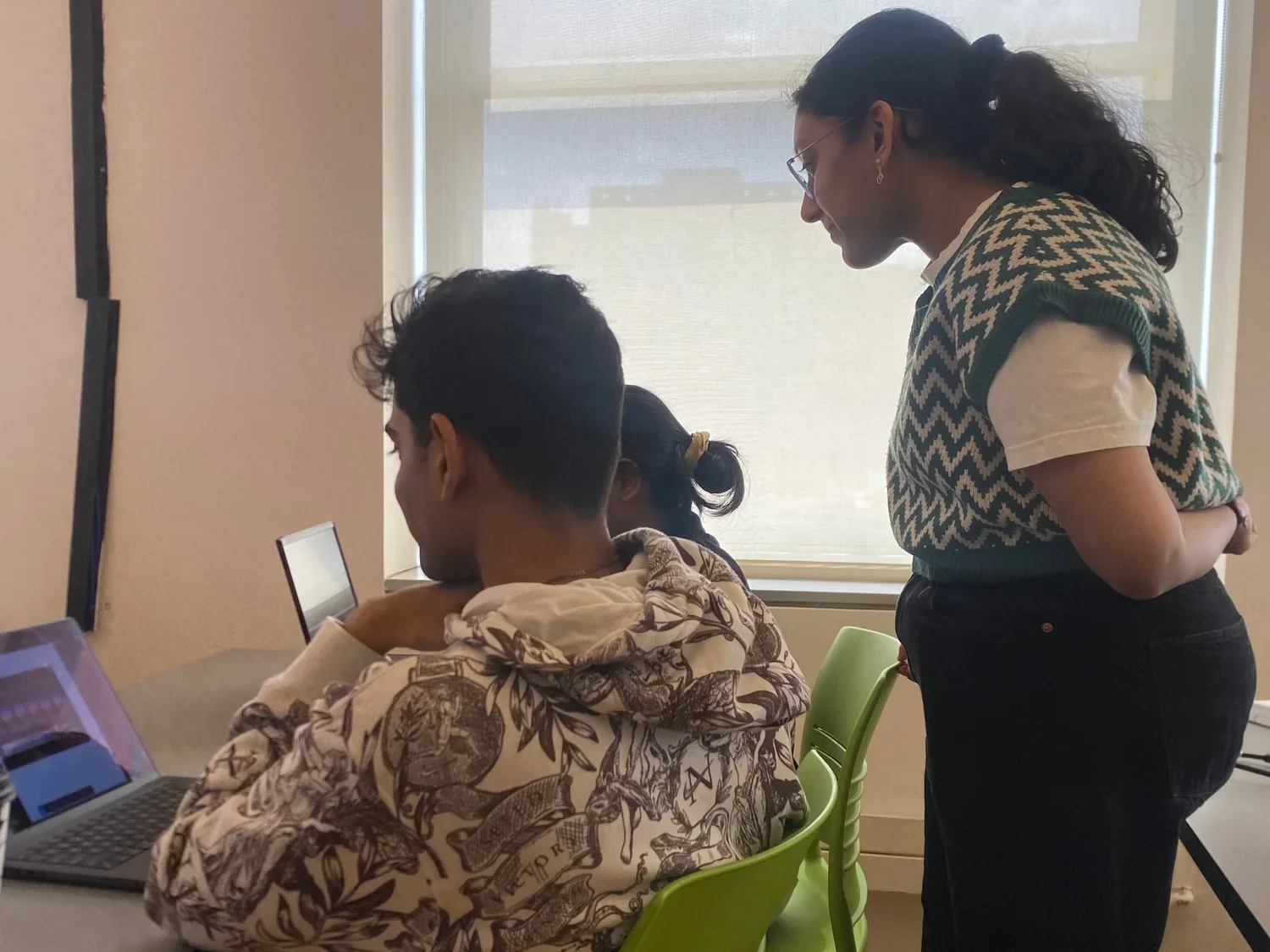 A woman is standing and leaning over a seated young man working on a laptop, with a young girl in the middle looking at a screen. They are in a room with a window and light-colored walls.