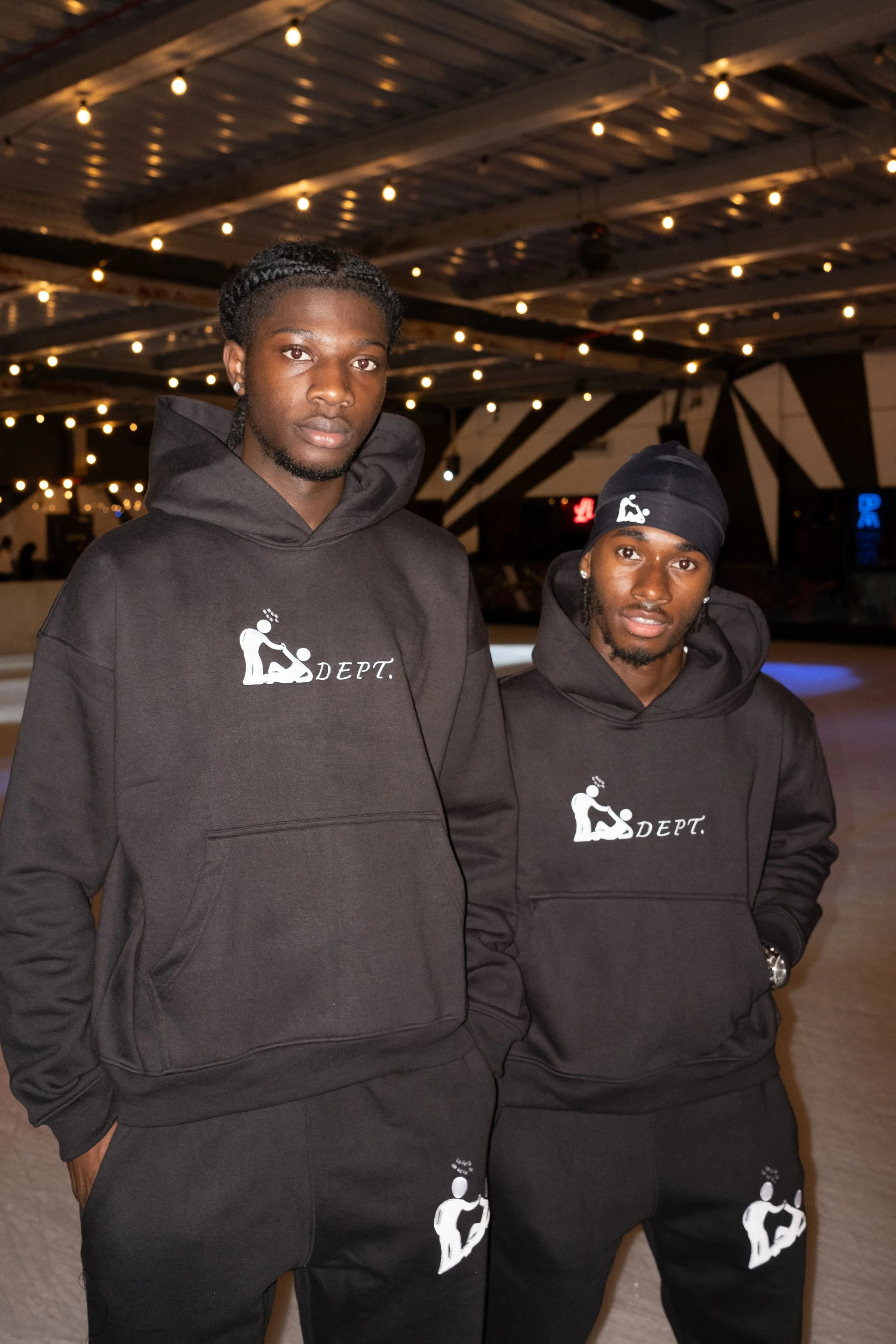 Two young men standing in an ice skating rink at night, both wearing black hoodies with a logo and the word 'DEPT' on them. The background is decorated with string lights and a wooden ceiling.