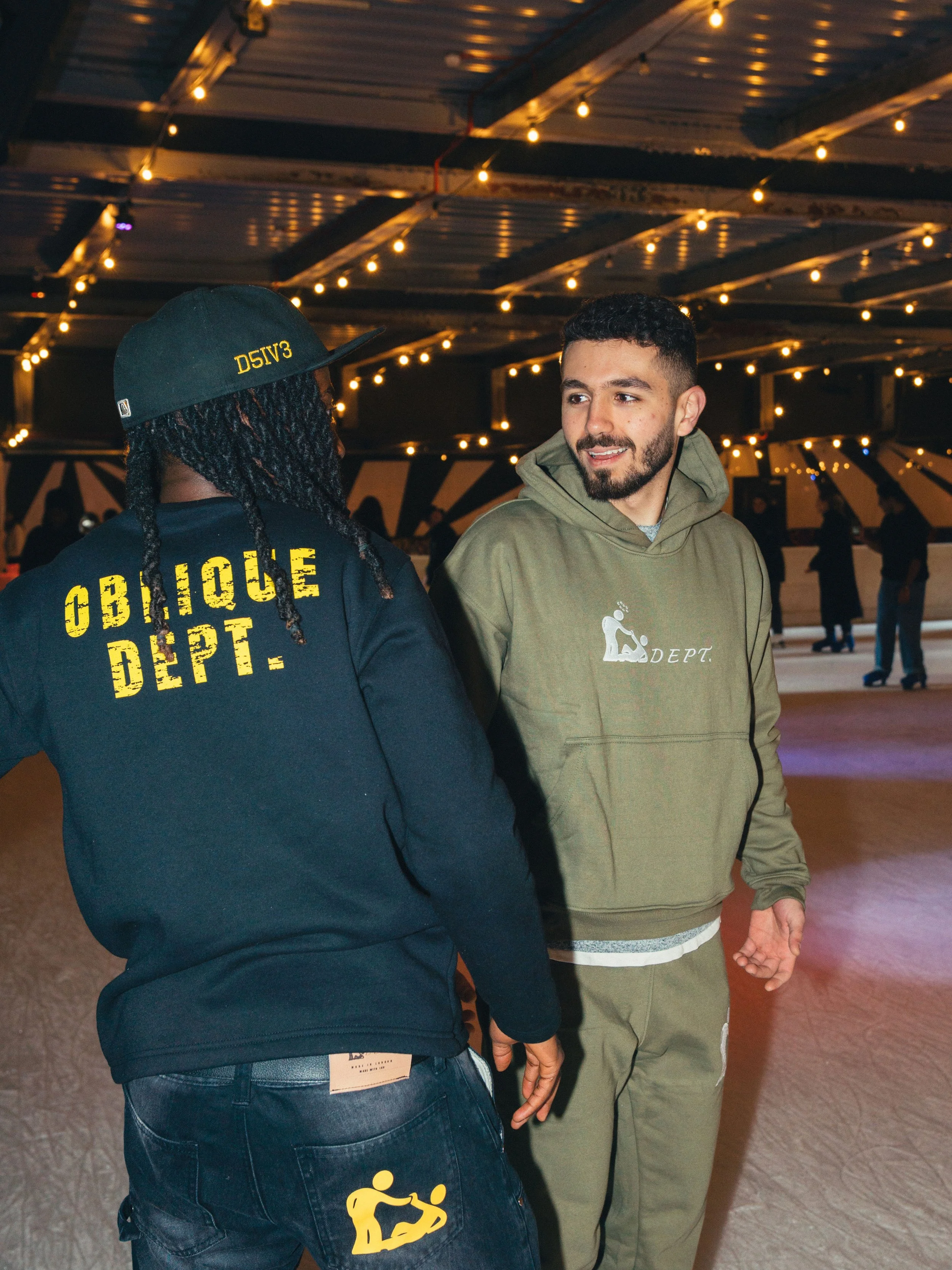 Two people at an indoor ice skating rink, talking and smiling. One is wearing a black hoodie with yellow text and a cap, the other is wearing a green hoodie and matching sweatpants.