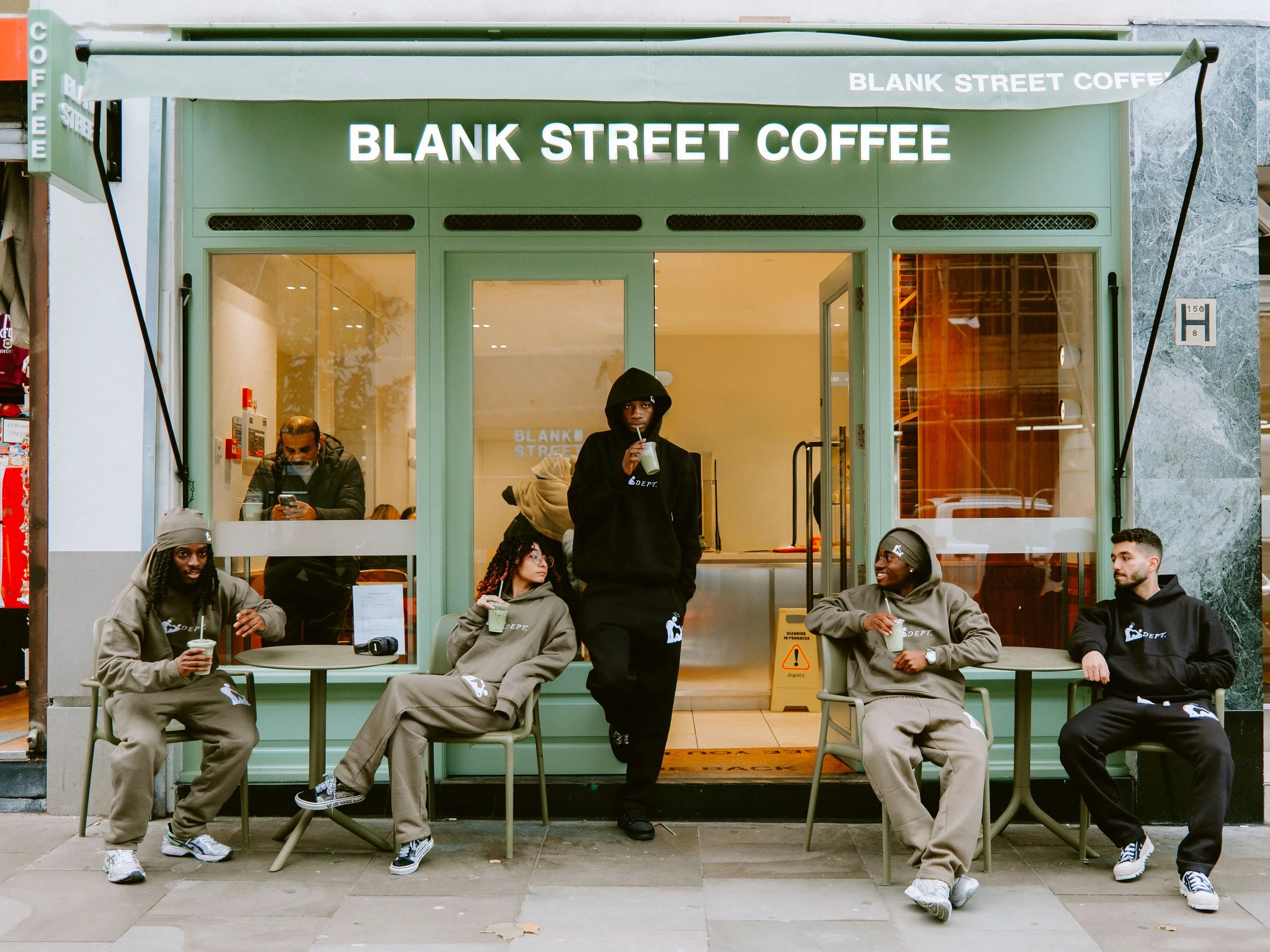 Five young people in hoodies and sweatpants sitting outside a coffee shop called 'Blank Street Coffee,' with one person standing and drinking from a cup.