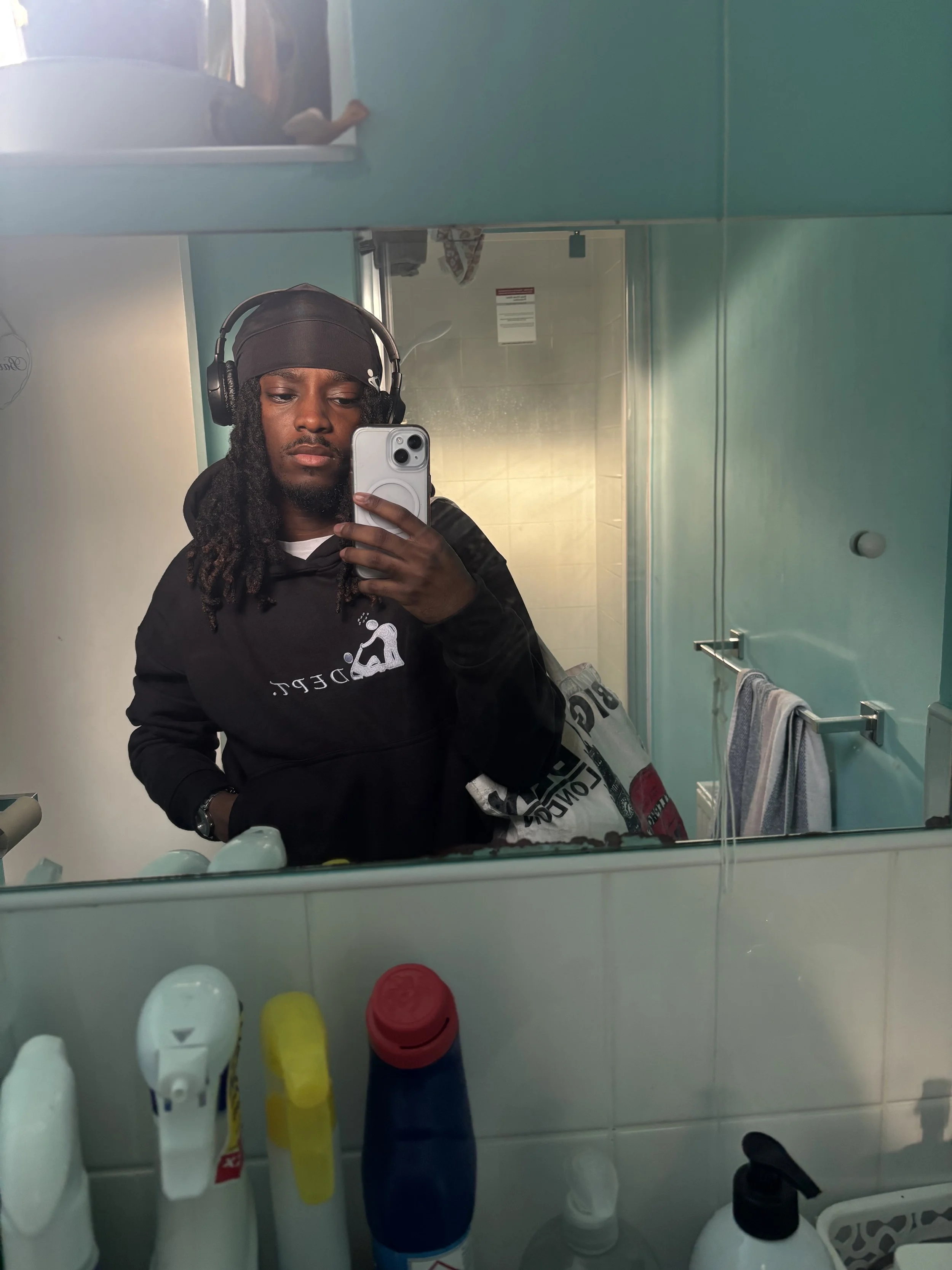 A man wearing a black hoodie, black headscarf, and large headphones taking a selfie in a bathroom mirror. He is holding a phone with a case. The bathroom has light green walls, a towel rack with a towel, and various cleaning products on the sink.