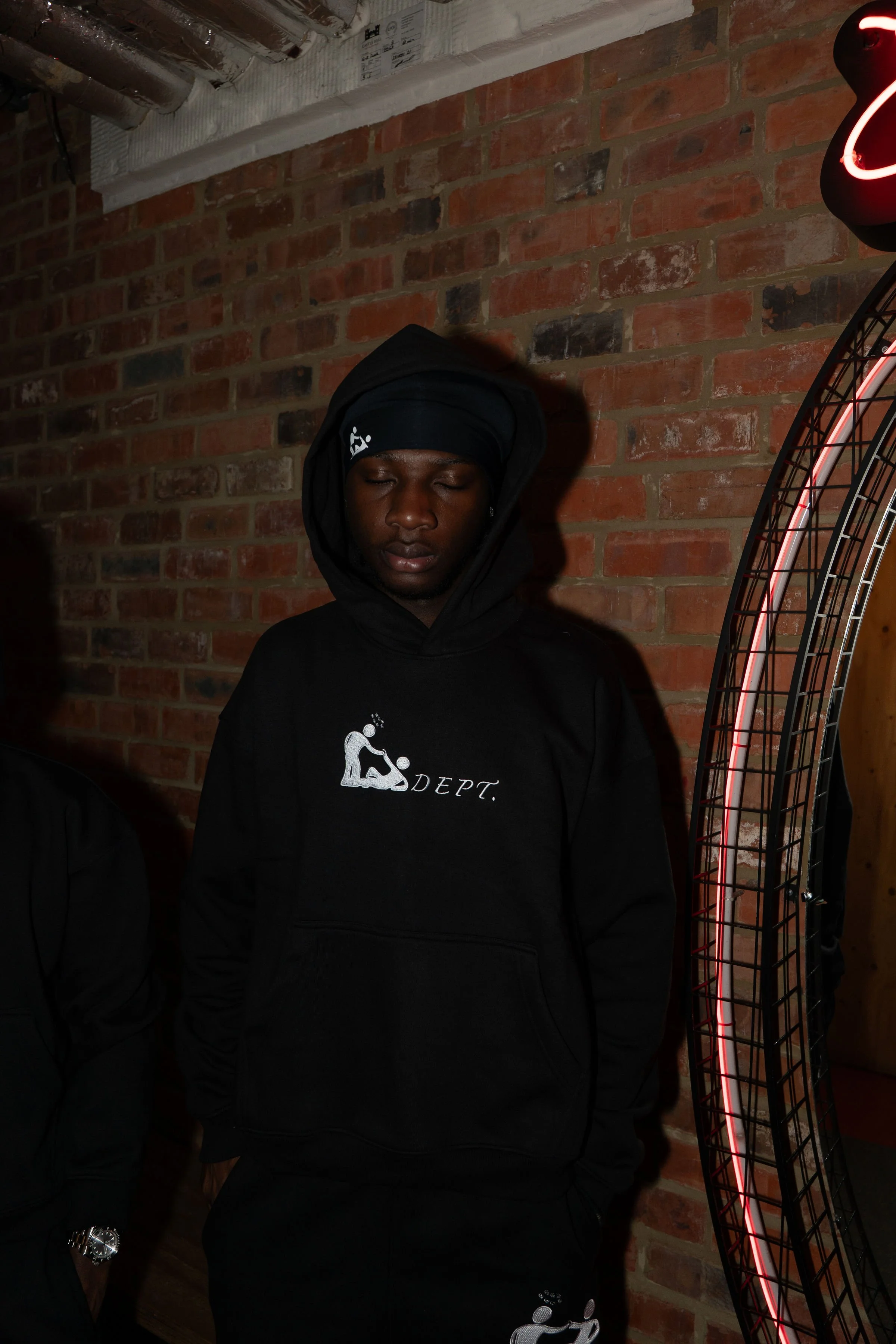 A man wearing a black hoodie with a graphic of a person and the word 'DEPT.' stands with eyes closed, in front of a brick wall, illuminated by dim lighting and a circular neon sign on the right.