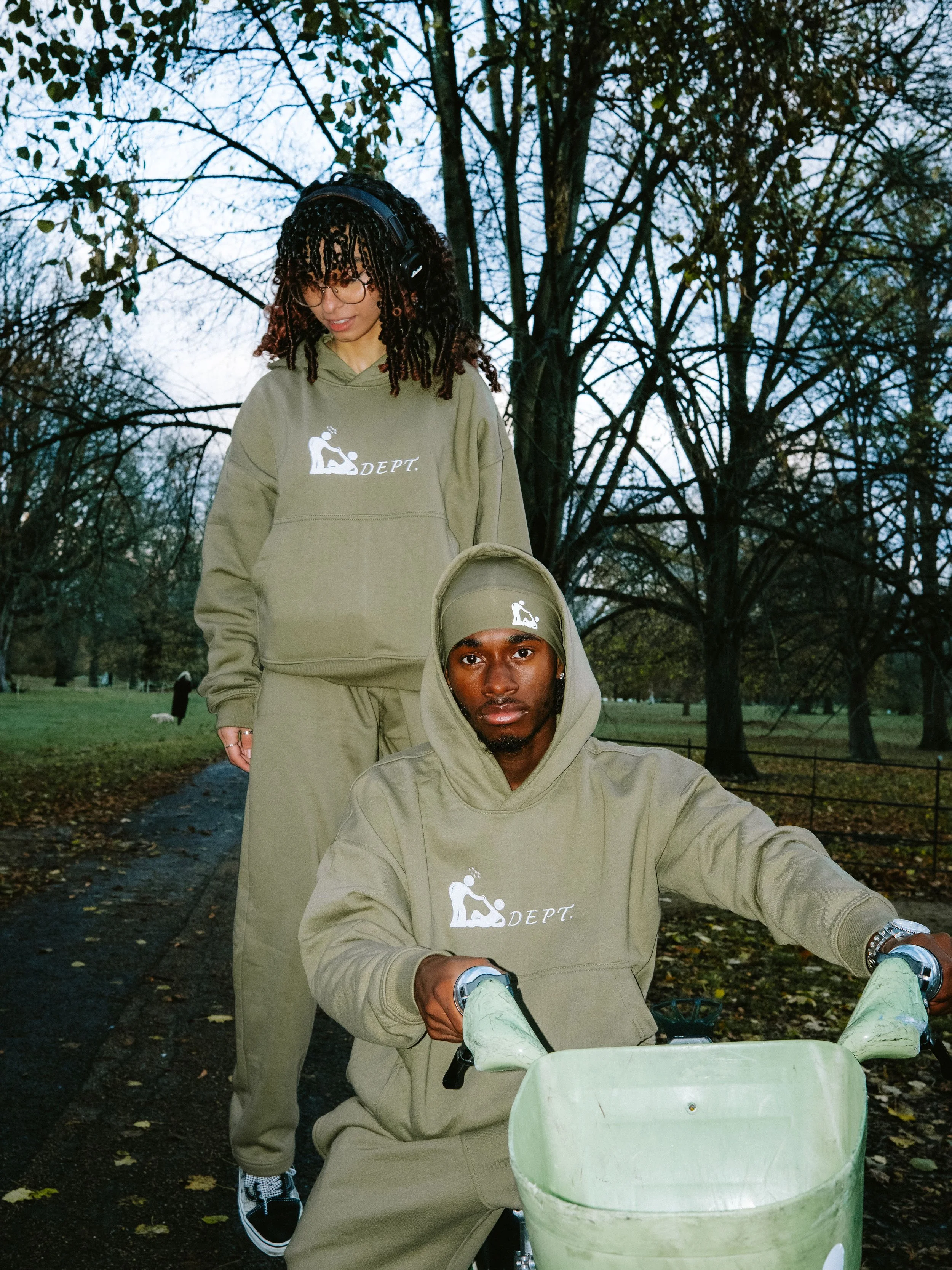 Two people wearing matching olive-green hoodies and pants with a logo; one sits on a bike with a basket, and the other stands behind on a park pathway with trees in the background.