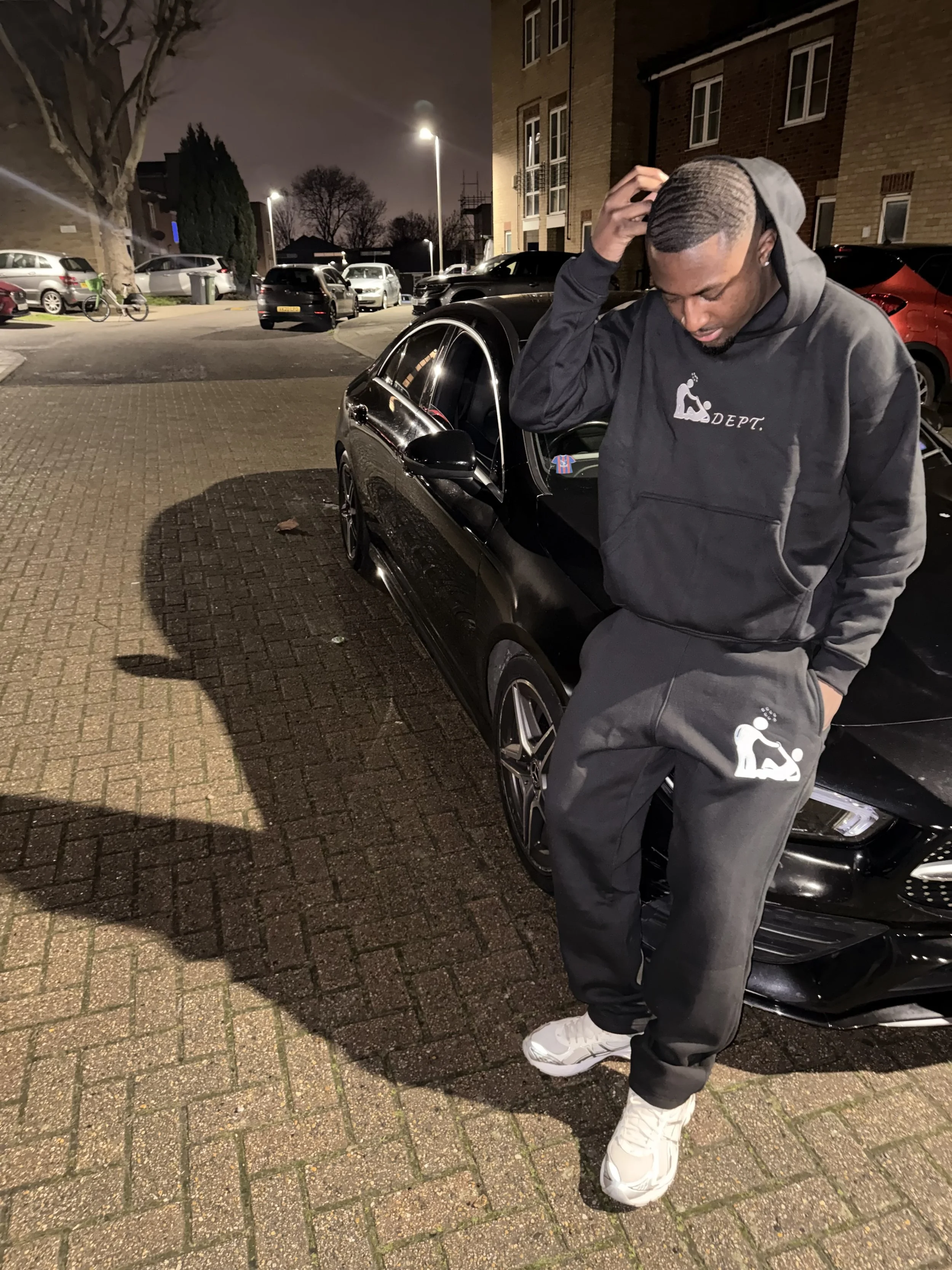 A man dressed in black athletic clothing with a hoodie and sweatpants leaning against a black car parked on a brick-paved street at night, looking down and scratching his head.