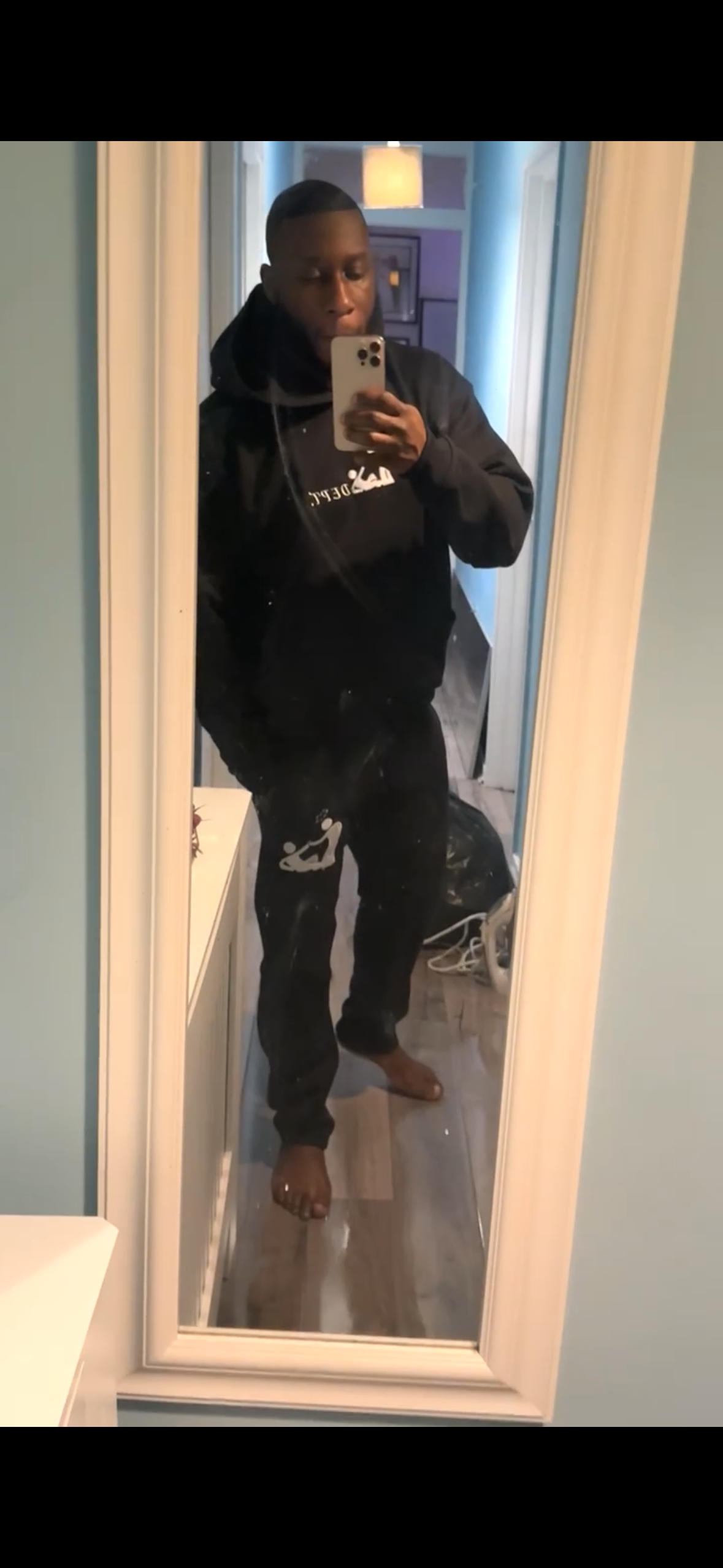 A person taking a mirror selfie in a hallway, wearing a black hoodie and black pants, standing barefoot on a wooden floor.