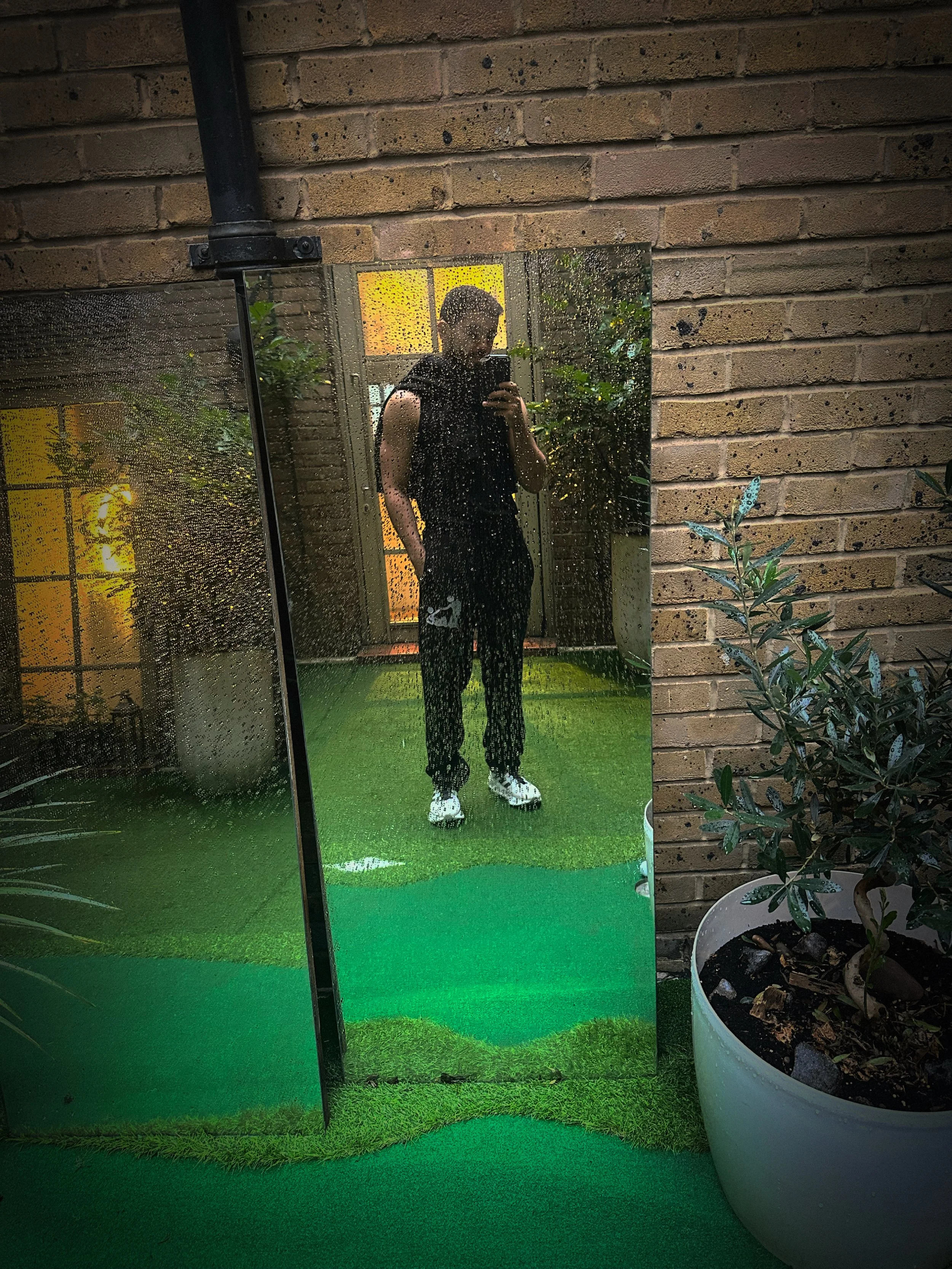 A person taking a mirror selfie outdoors on a rainy day, with a brick wall and green plants in the background.