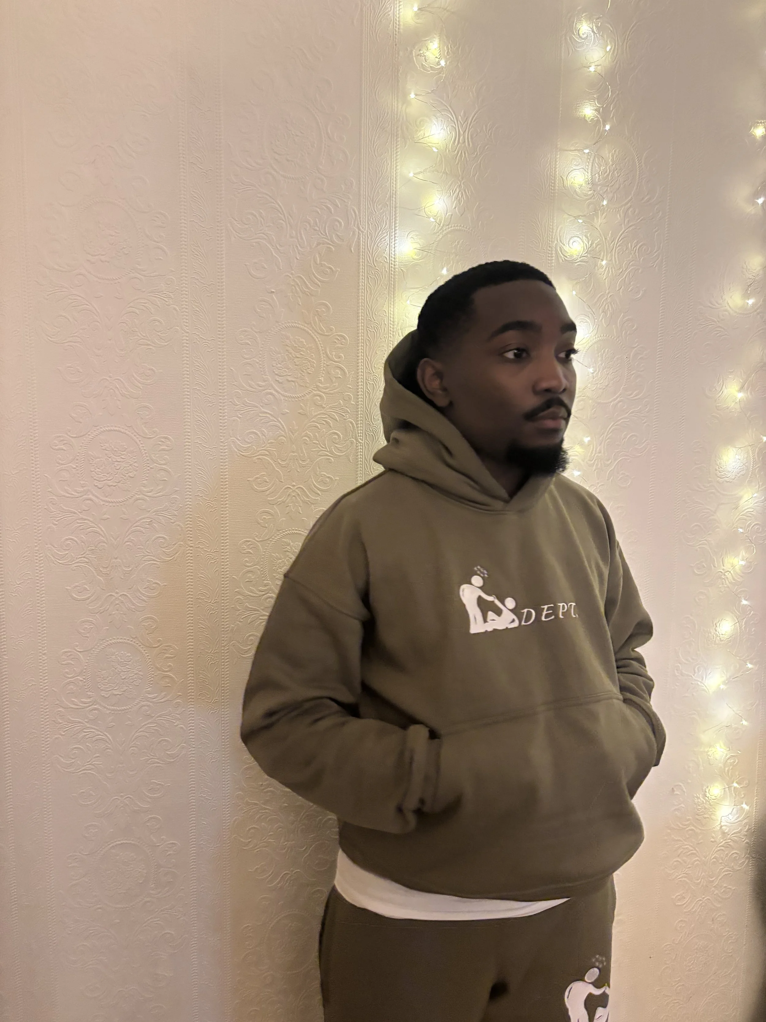 A young man in a brown hoodie with a graphic of a baseball player and the text 'D EPT' stands against a beige wall decorated with string lights.