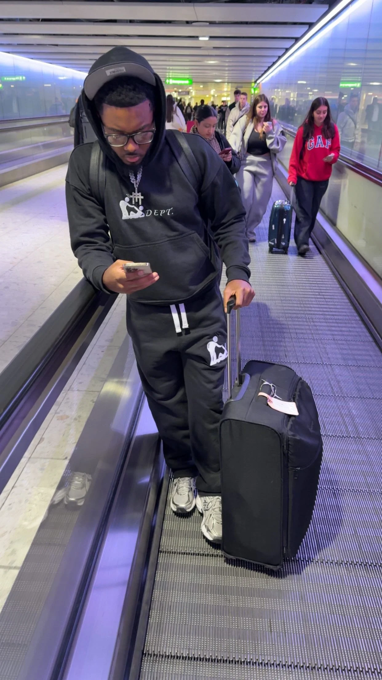A young man wearing glasses, a black hoodie, and black sweatpants with white sneakers is standing on a moving walkway at an airport, looking at his phone. He has a black rolling suitcase next to him. In the background, several people are walking or s