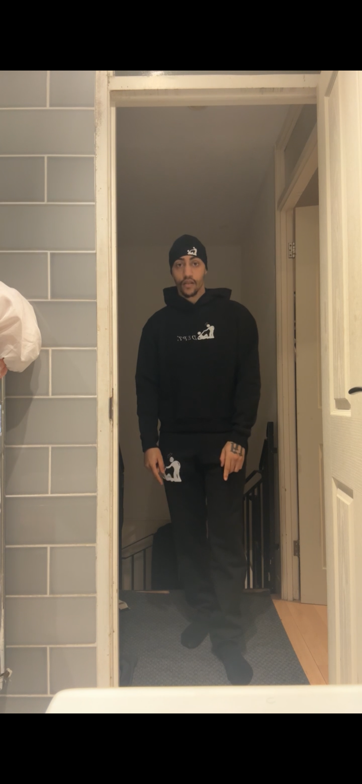 A man wearing a black hoodie, black pants, and a black beanie, standing in a doorway and taking a mirror selfie in a dimly lit room.