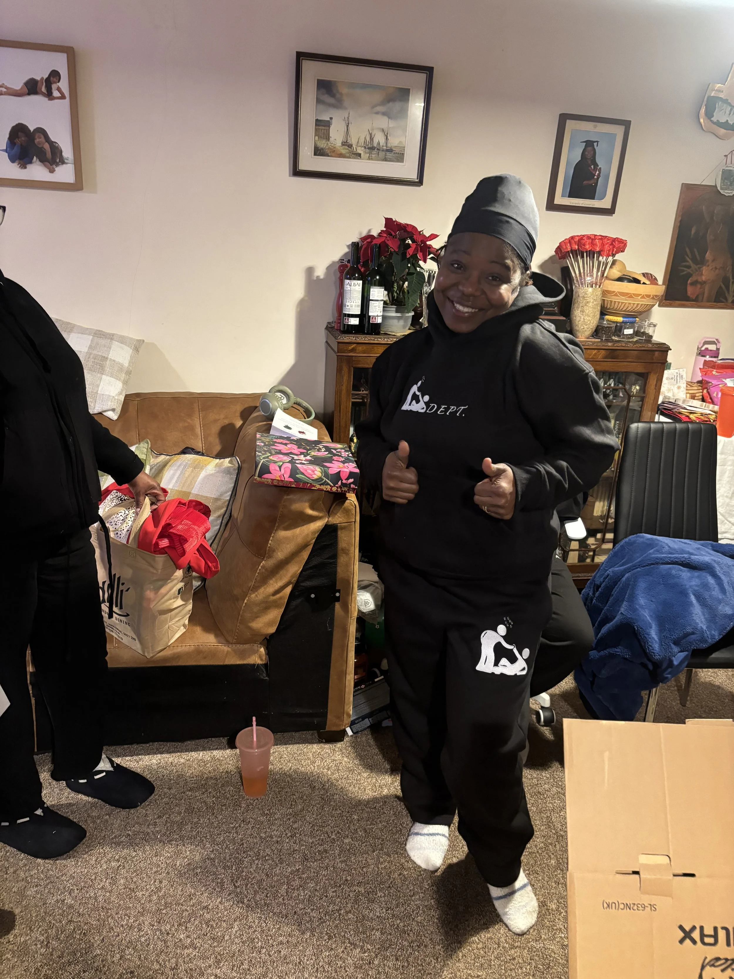 A woman wearing a black hoodie and matching pants, smiling and giving two thumbs up in a living room decorated with framed pictures and holiday decorations.