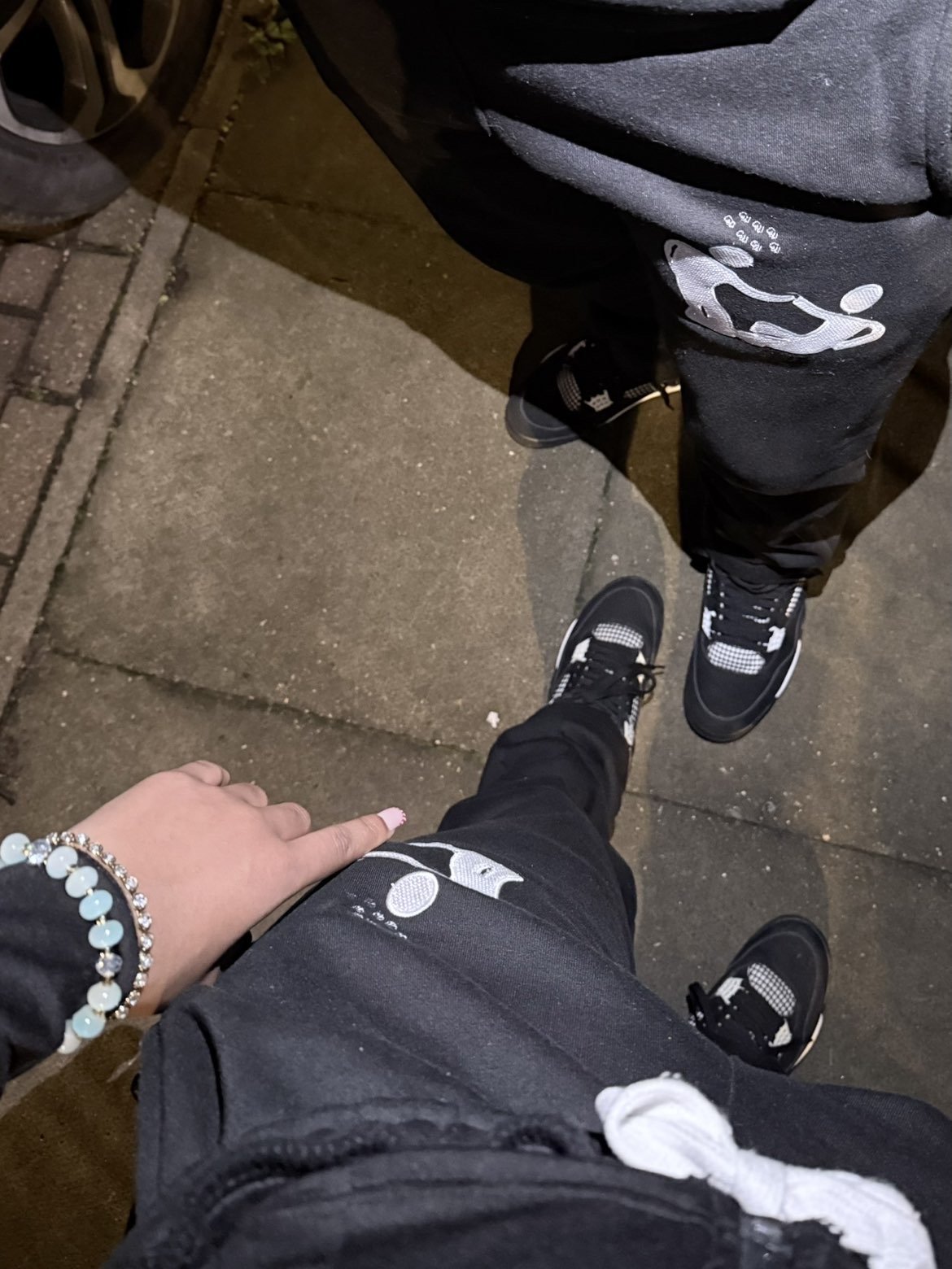 Top-down view of two people standing on a sidewalk, wearing black sneakers with white accents, black pants, and sweatshirts. One person's hand is visible, wearing a light blue bracelet.