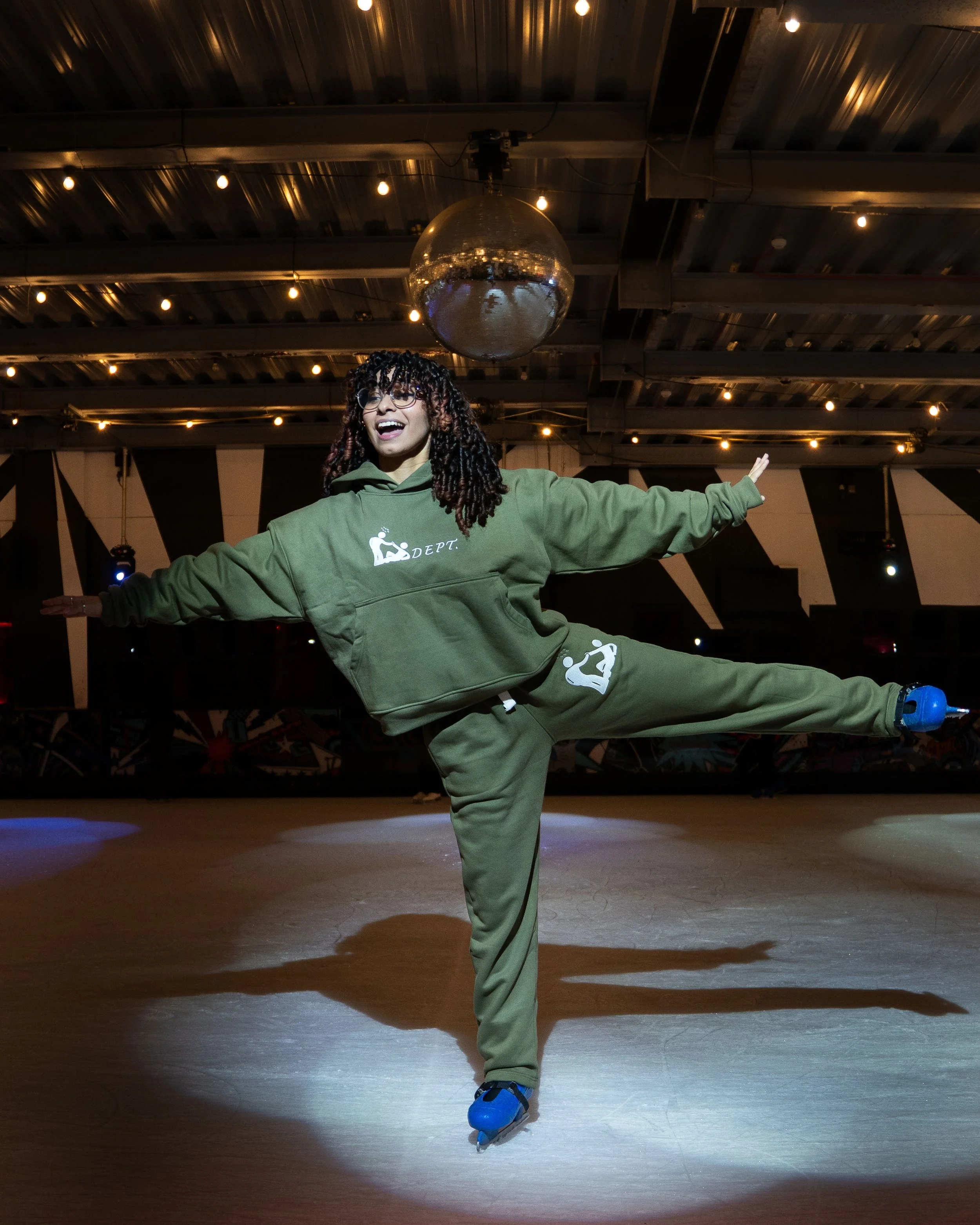 A woman roller skating indoors, balancing on one leg with arms extended, wearing green sportswear and glasses, under warm lighting with a disco ball overhead.