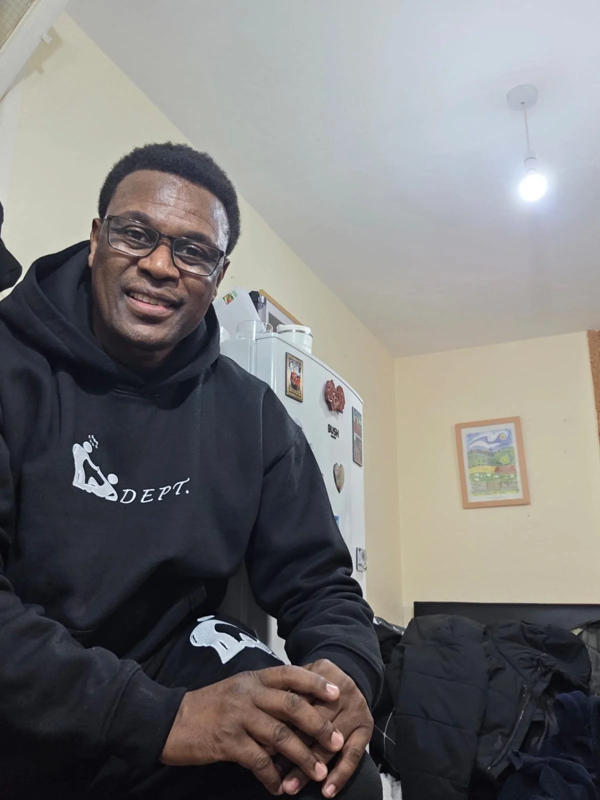 A man sitting at a table in a kitchen, smiling, wearing a black hoodie and glasses.