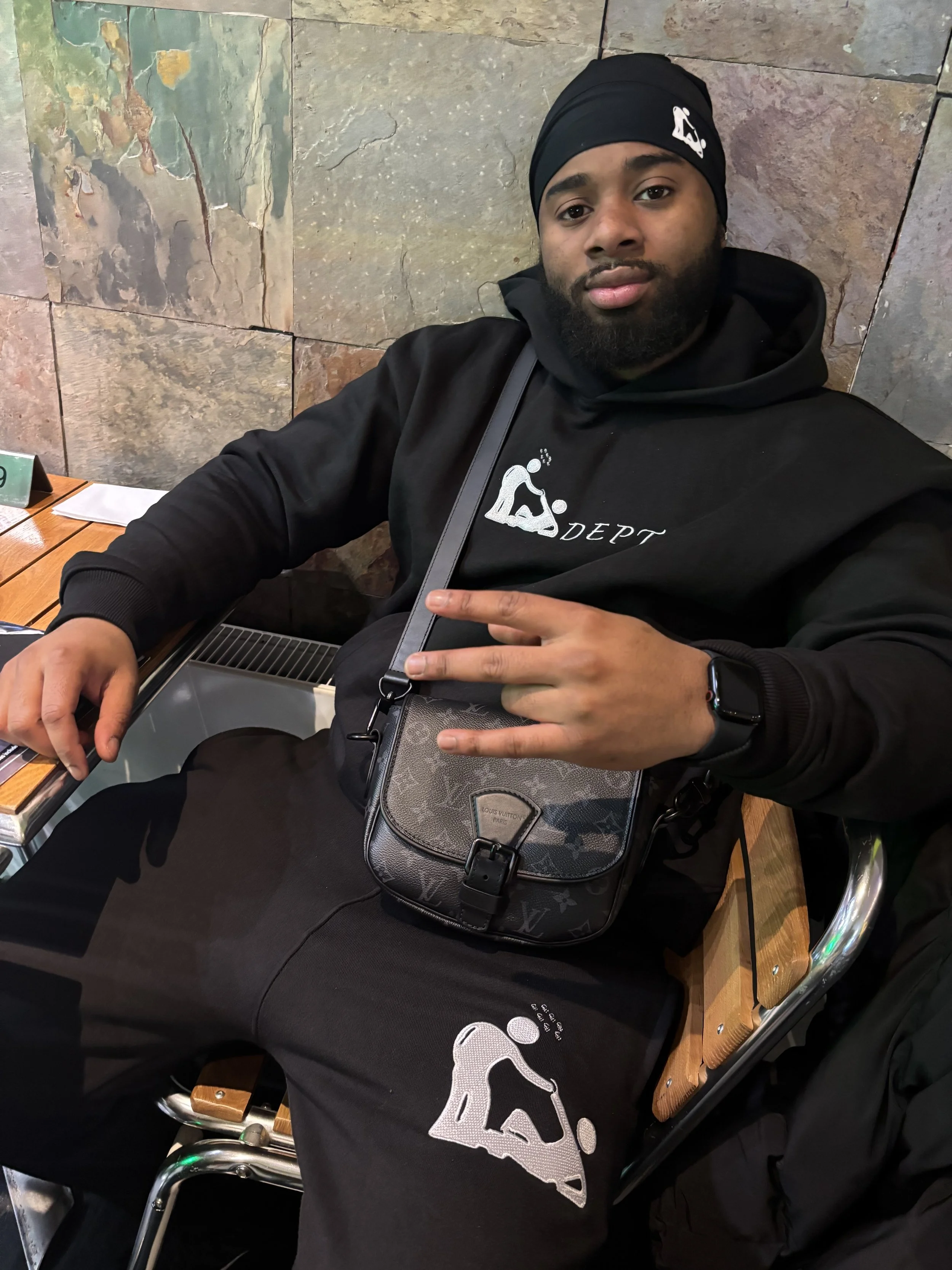A man sitting on a wooden bench at a cafe, wearing a black hoodie, black sweatpants, and a black head wrap with a logo. He has a crossbody bag and is making a peace sign with his right hand. The background features a stone wall.