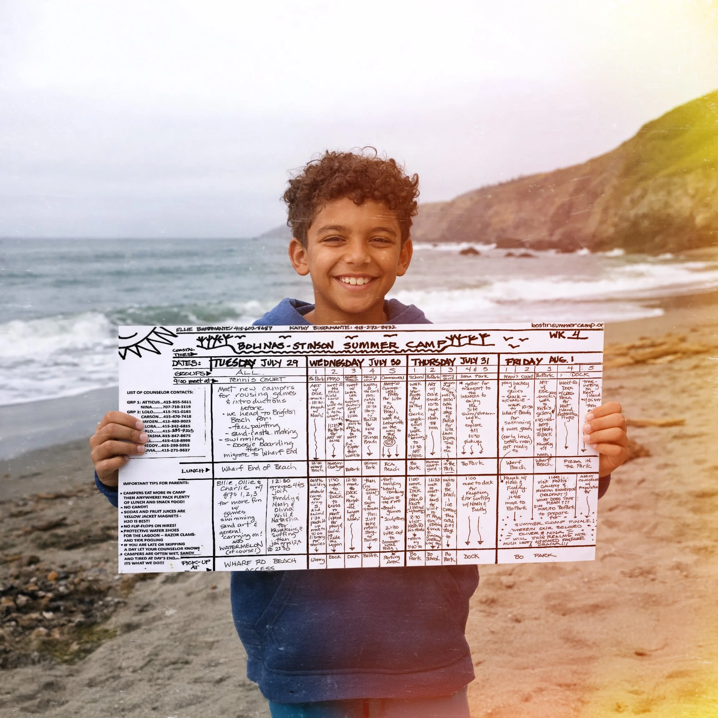 A smiling boy holding a weekly schedule at the beach with ocean and cliffs in the background.