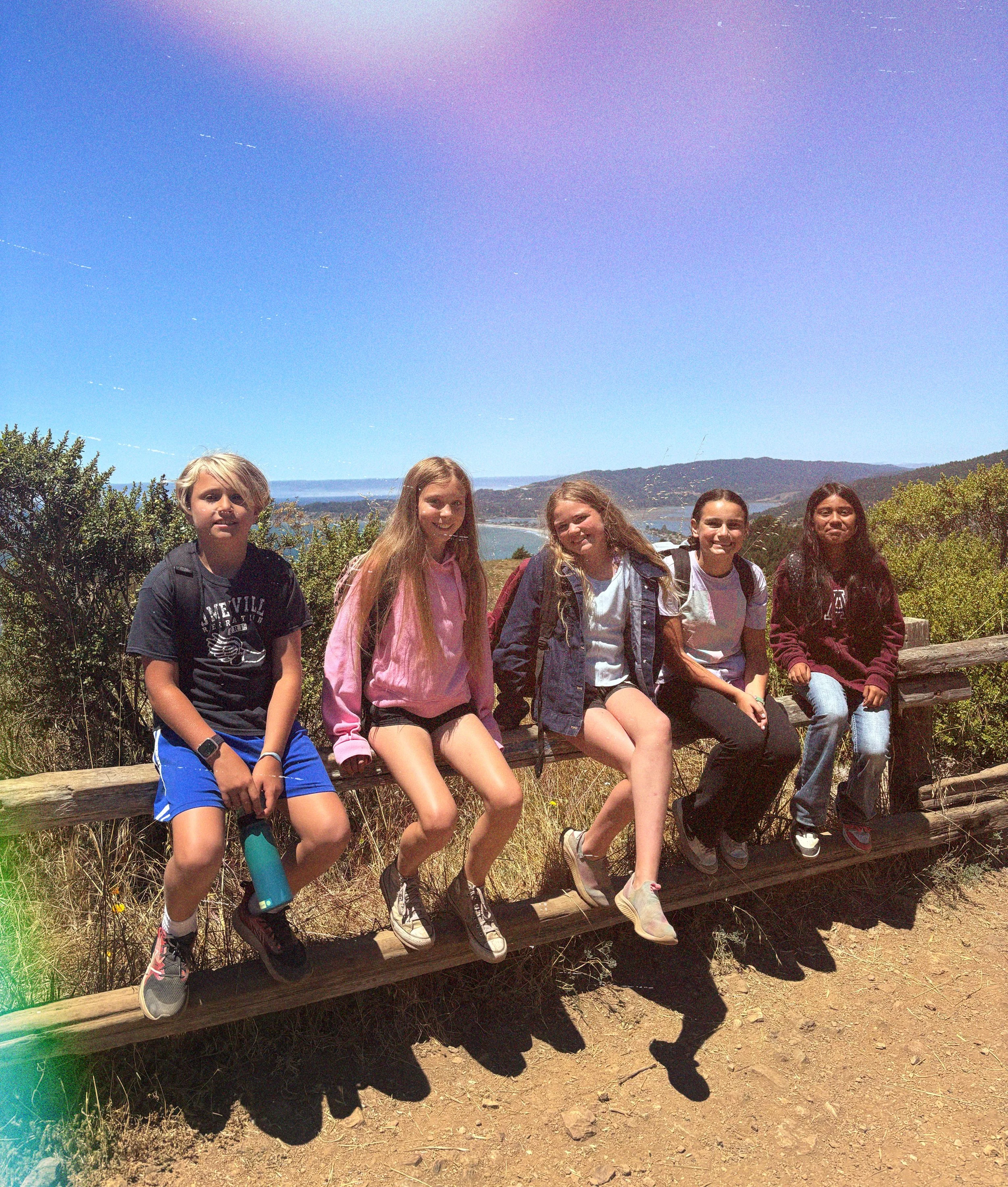 Group of five young girls sitting on a wooden bench outdoors with a scenic background of trees, water, and hills under a clear blue sky.