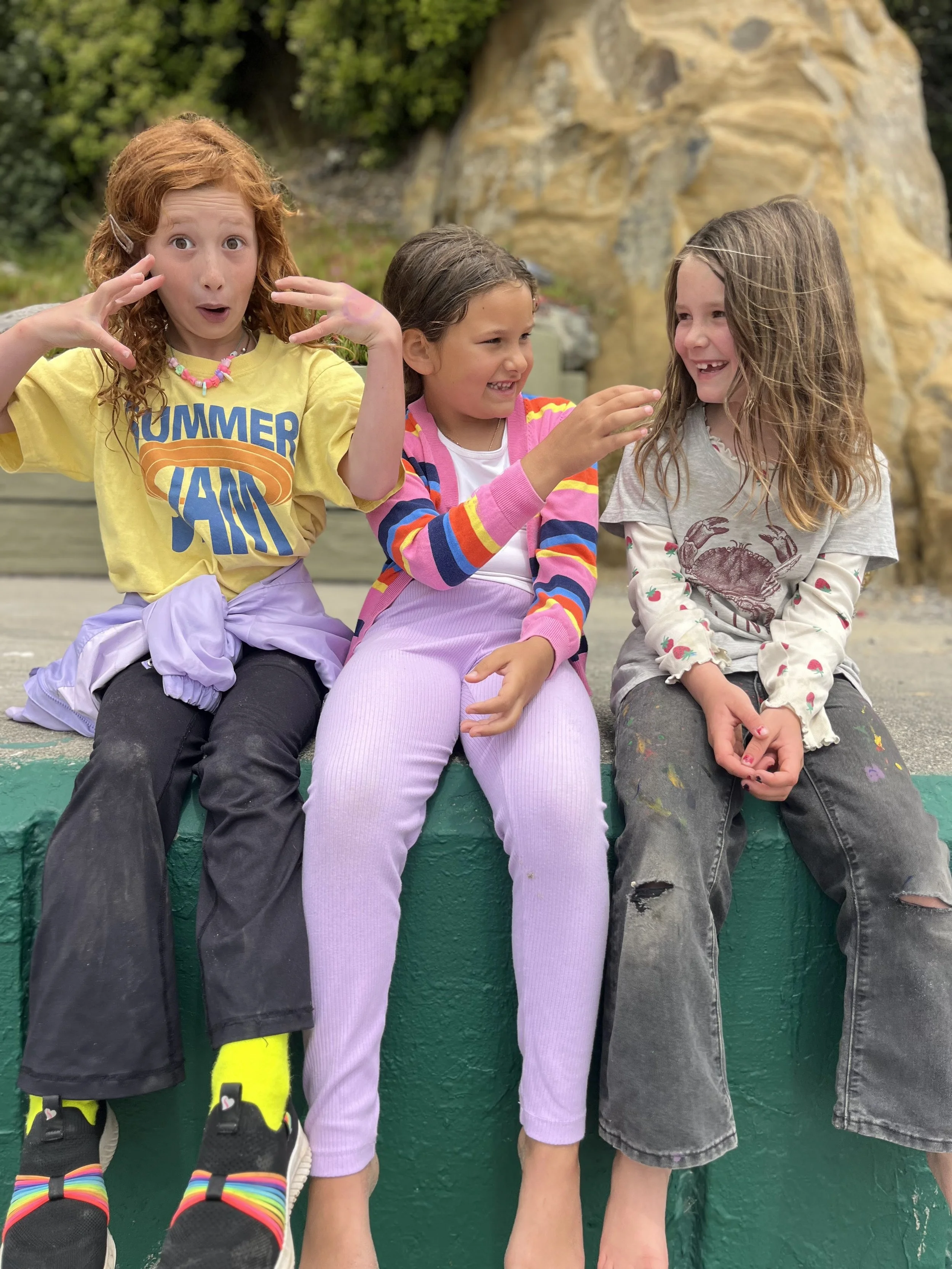 Three young girls sitting on a green ledge, making funny faces and smiling, outdoors with rocks and greenery in the background.