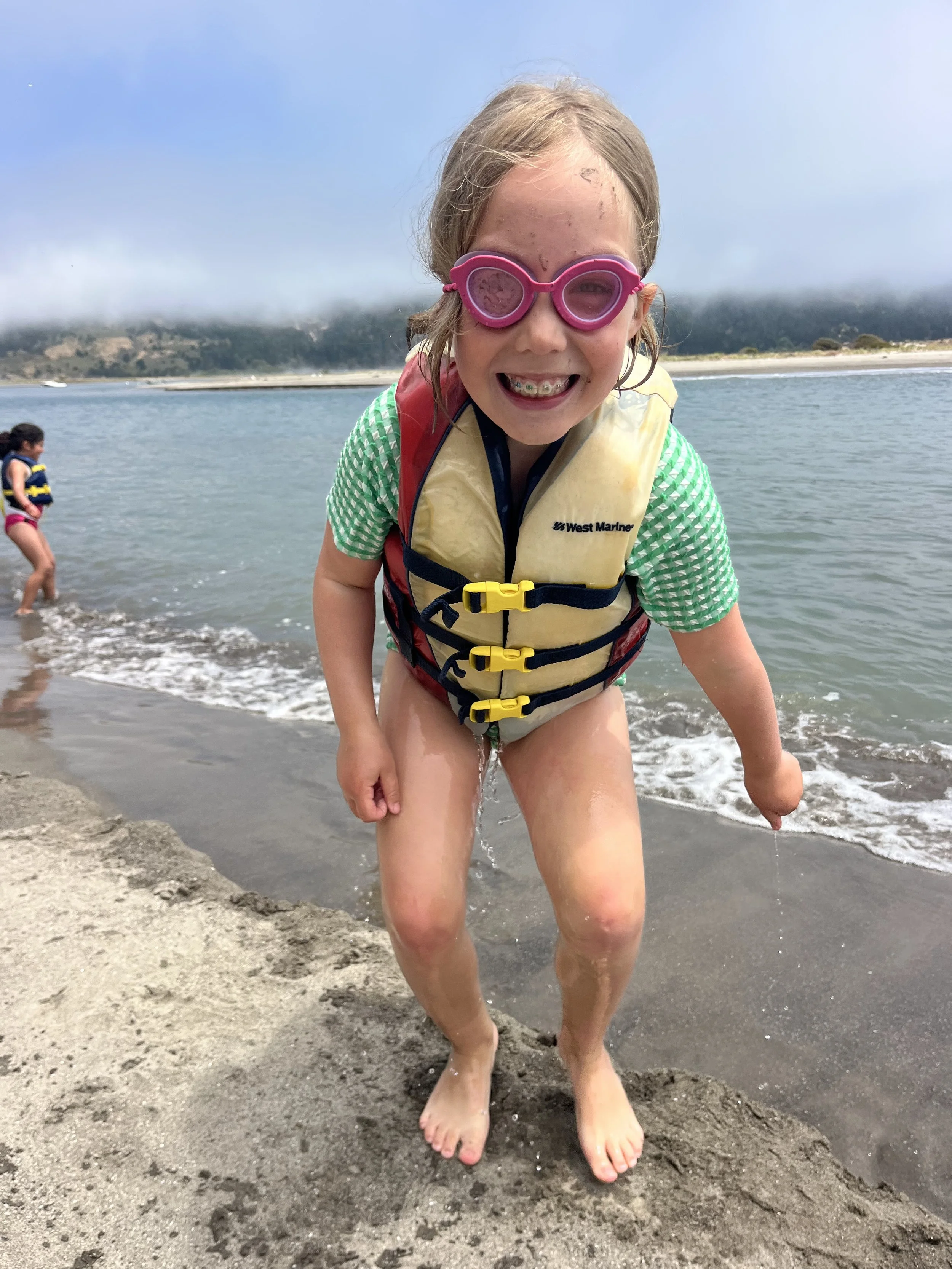 A young girl smiling at the camera while standing on a beach, wearing a life jacket and pink swim goggles, with another child in the background near the water.