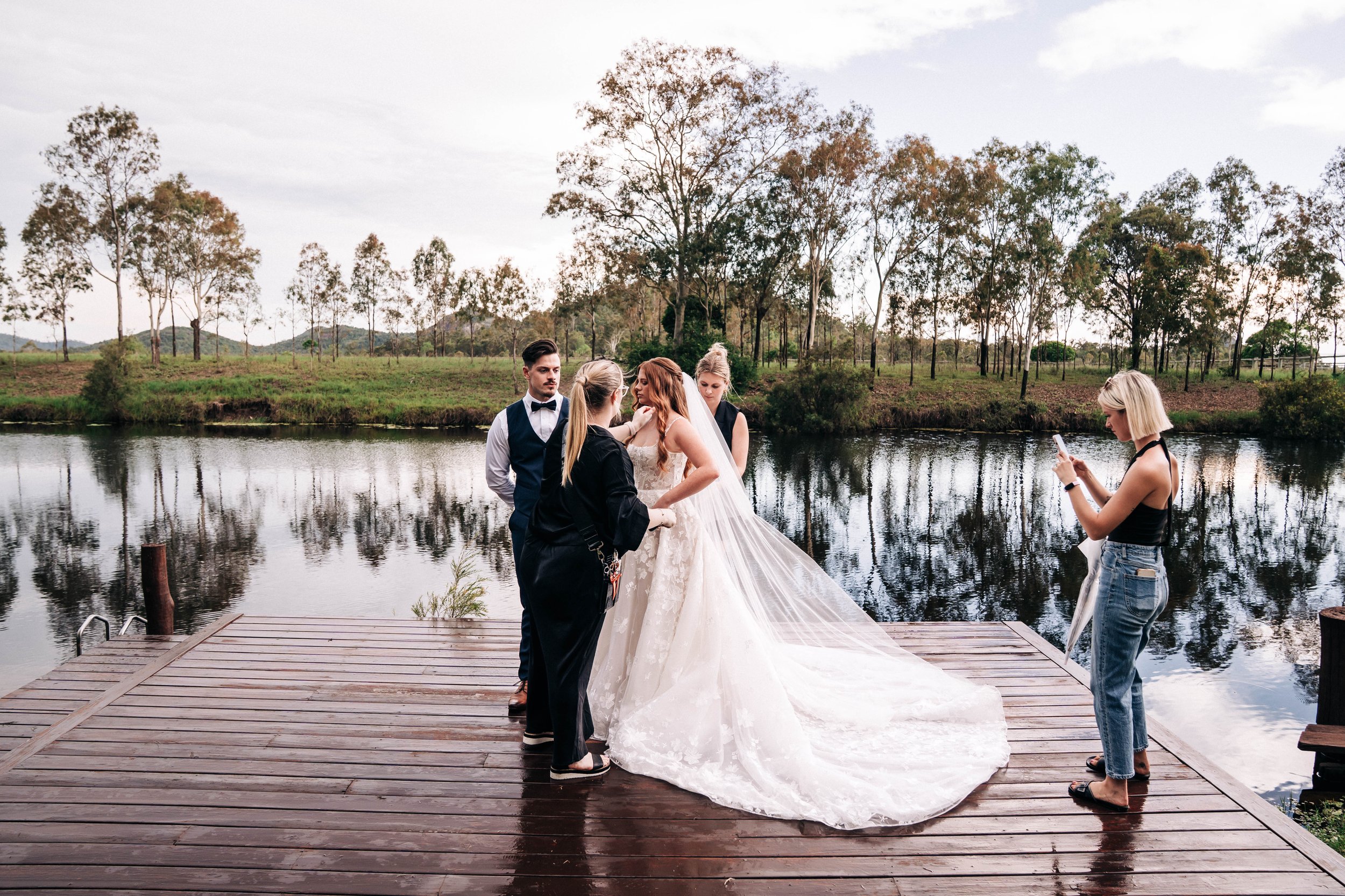 A bride and groom along with two women are on a wooden dock by a river, with trees and water reflections in the background, during a wedding photoshoot.