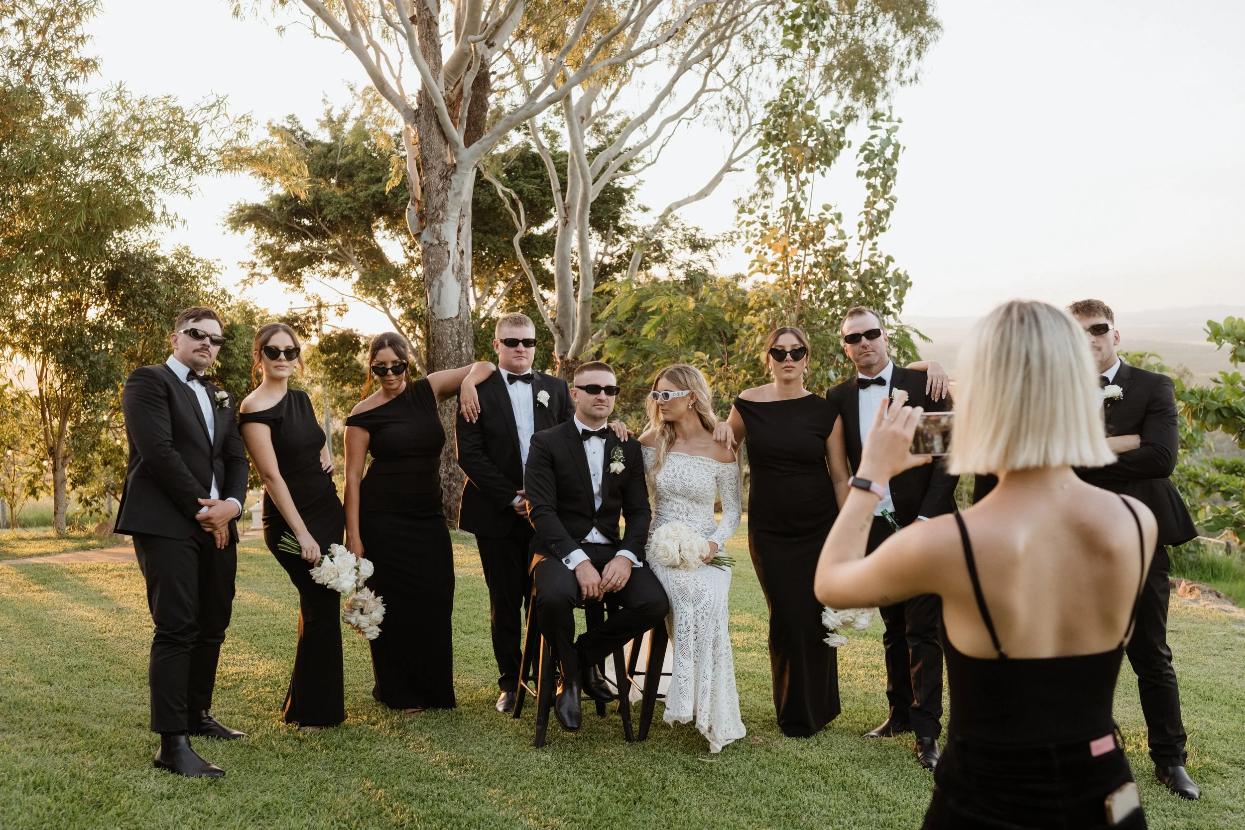 Group of people in formal attire, wearing black sunglasses, posing outdoors during sunset, with a woman in a white lace dress seated in the center and a woman in black taking a photo in the foreground.