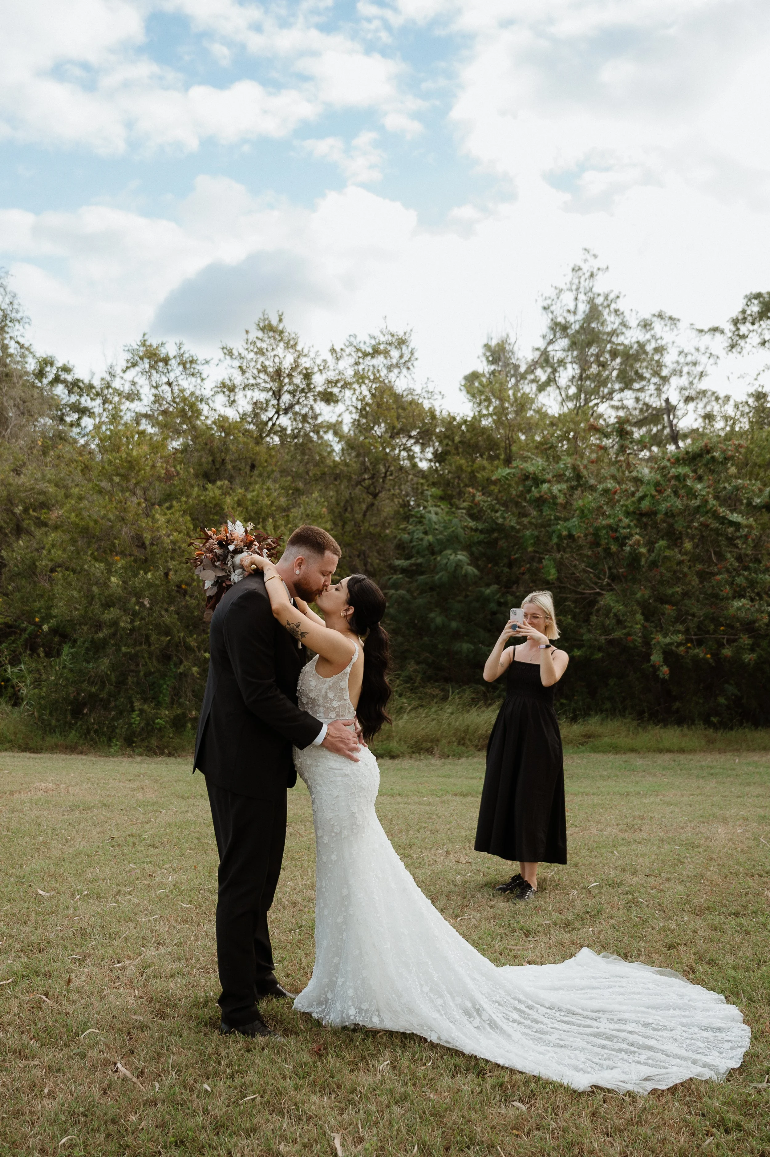 A bride and groom share a kiss outdoors during their wedding ceremony, with a woman in a black dress taking a photo nearby.