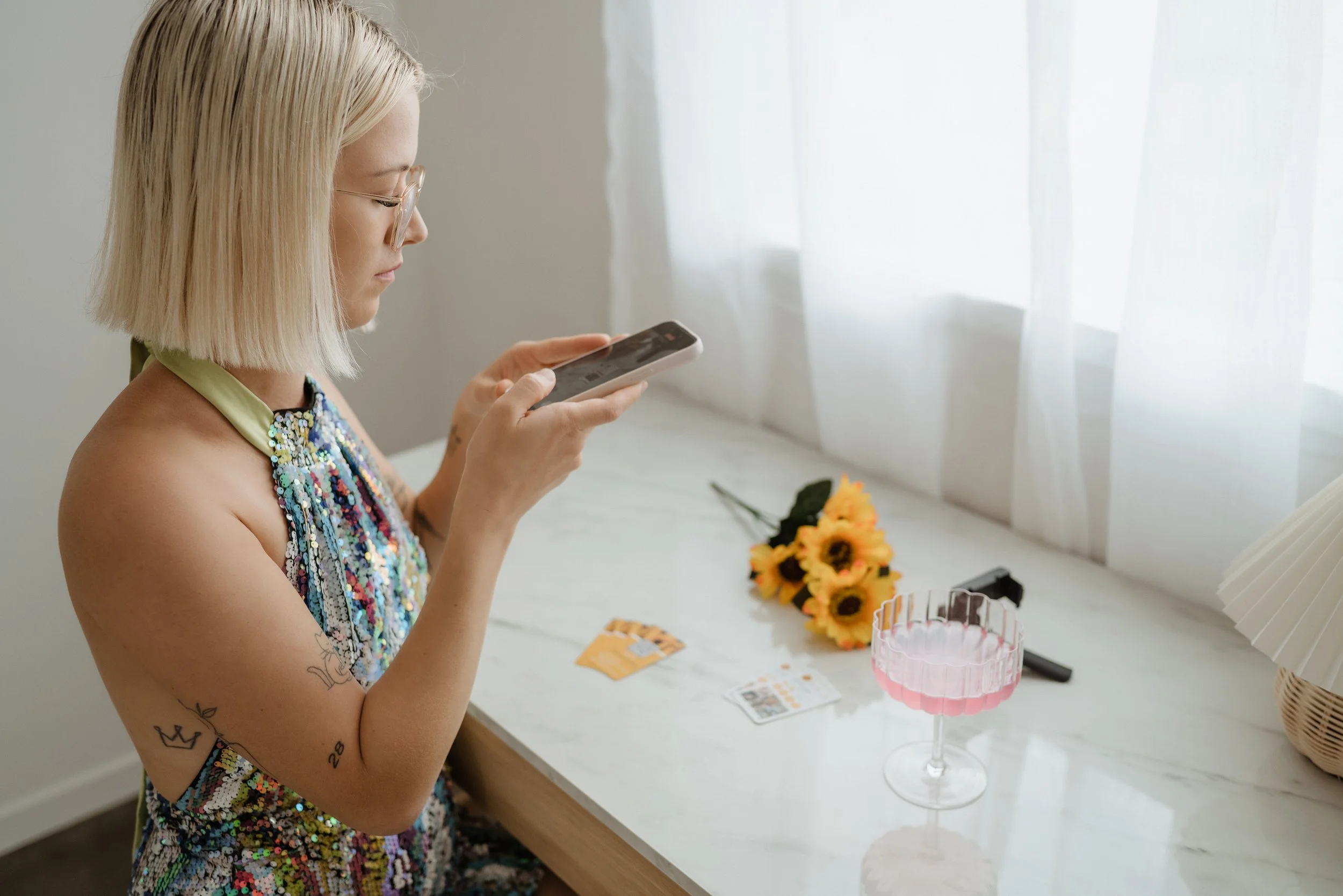 A woman with blonde hair, wearing glasses and a colorful sequin dress, sitting at a white table looking at her phone. On the table are a small bouquet of sunflowers, a pink glass dish, some papers, credit cards, and a pair of scissors, with white curtains in the background.