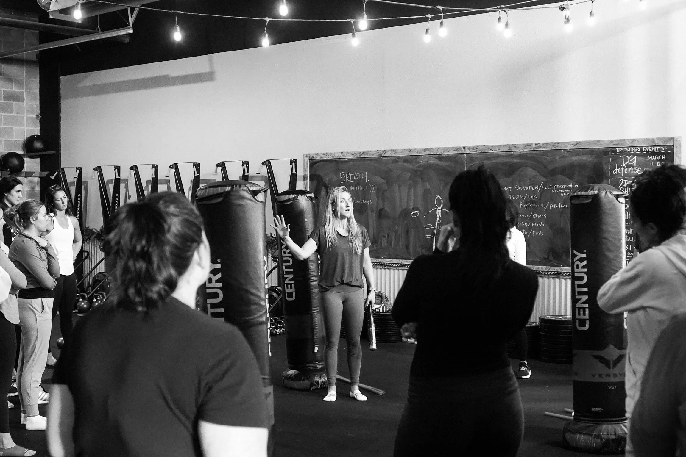Instructor leading a fitness class in a gym, with students standing around in front of punching bags, in a black-and-white photo.