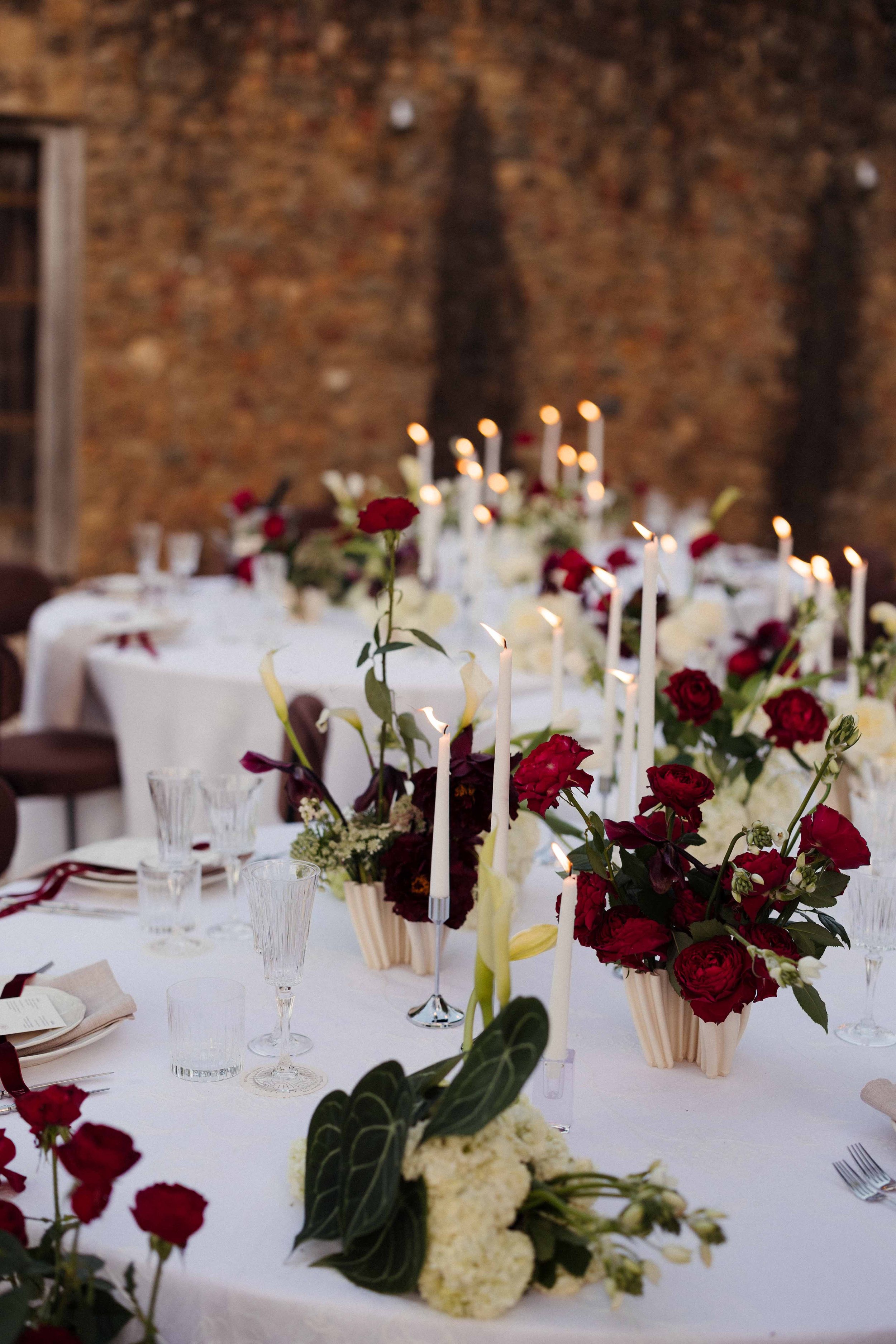 Elegant table setting with floral centerpieces, white candles, glassware, and white tablecloths at a formal event, against a brick wall background.