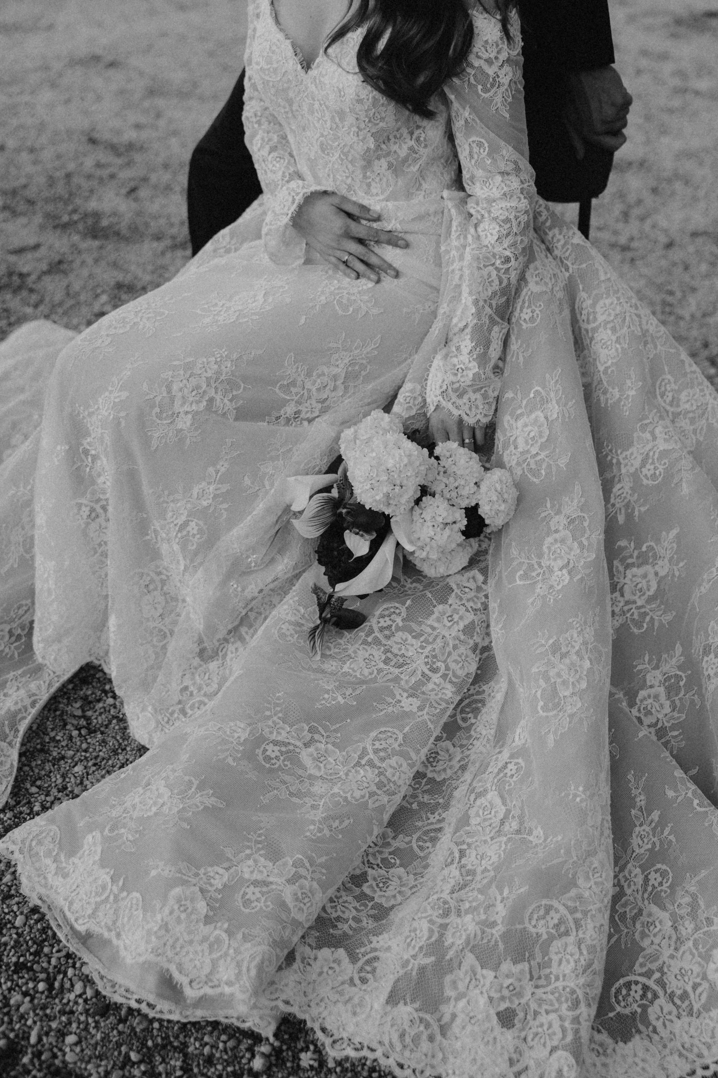 A black-and-white photograph of a bride in a lace wedding dress holding a bouquet of flowers, seated with a person behind her on a textured ground.