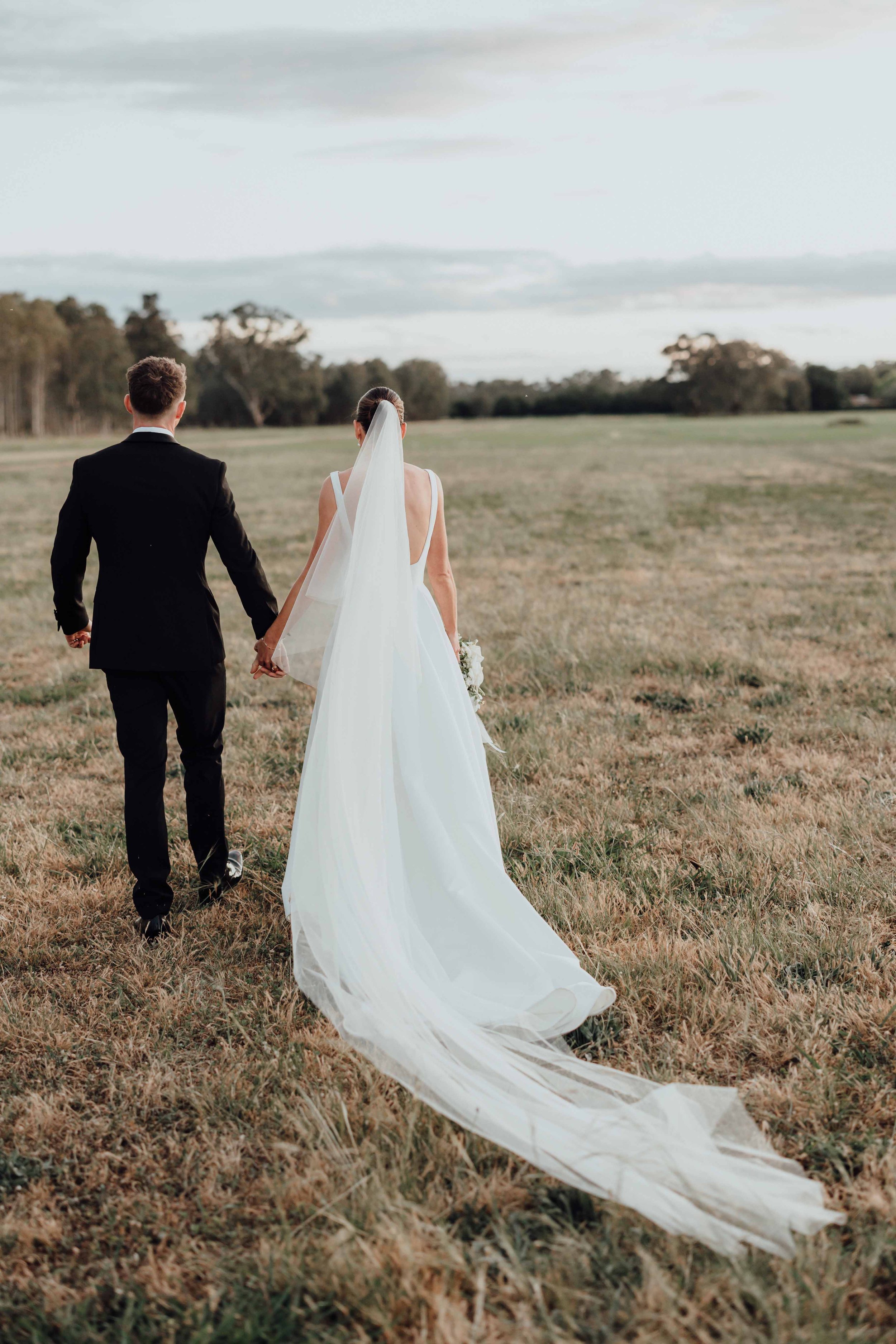 A bride in a white wedding dress and veil holding hands with a groom in a black suit walking across an open field.