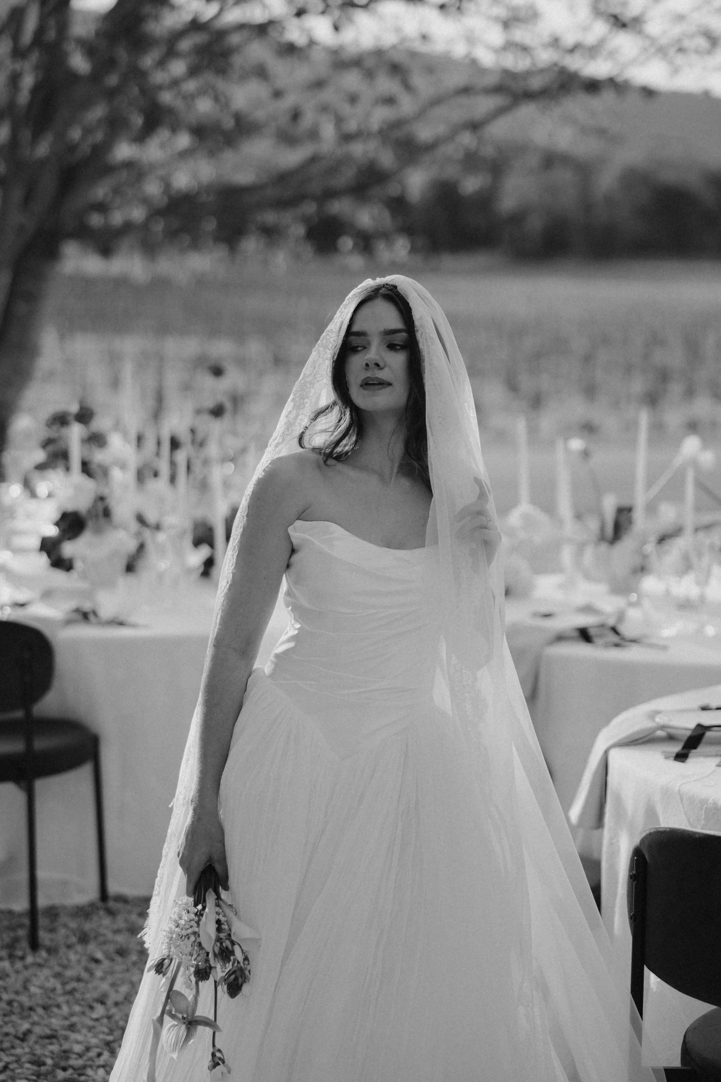 Black and white photo of a woman in a wedding dress holding a bouquet, standing outdoors under a tree, with a blurred background of a vineyard and outdoor tables.