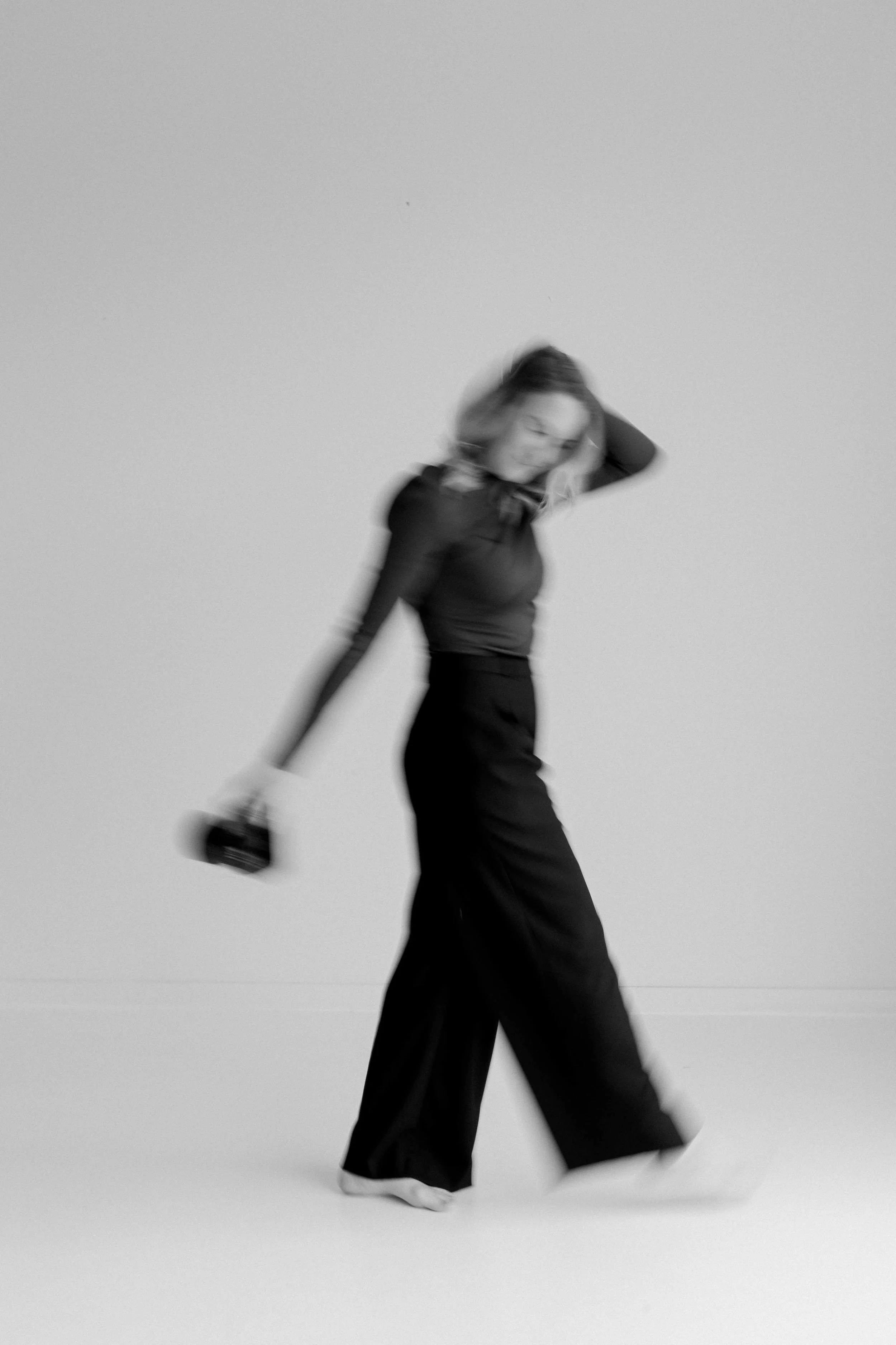 A woman in black pants and a black long-sleeve top walking barefoot on a plain background, holding a camera in her right hand, with a blurred motion effect.