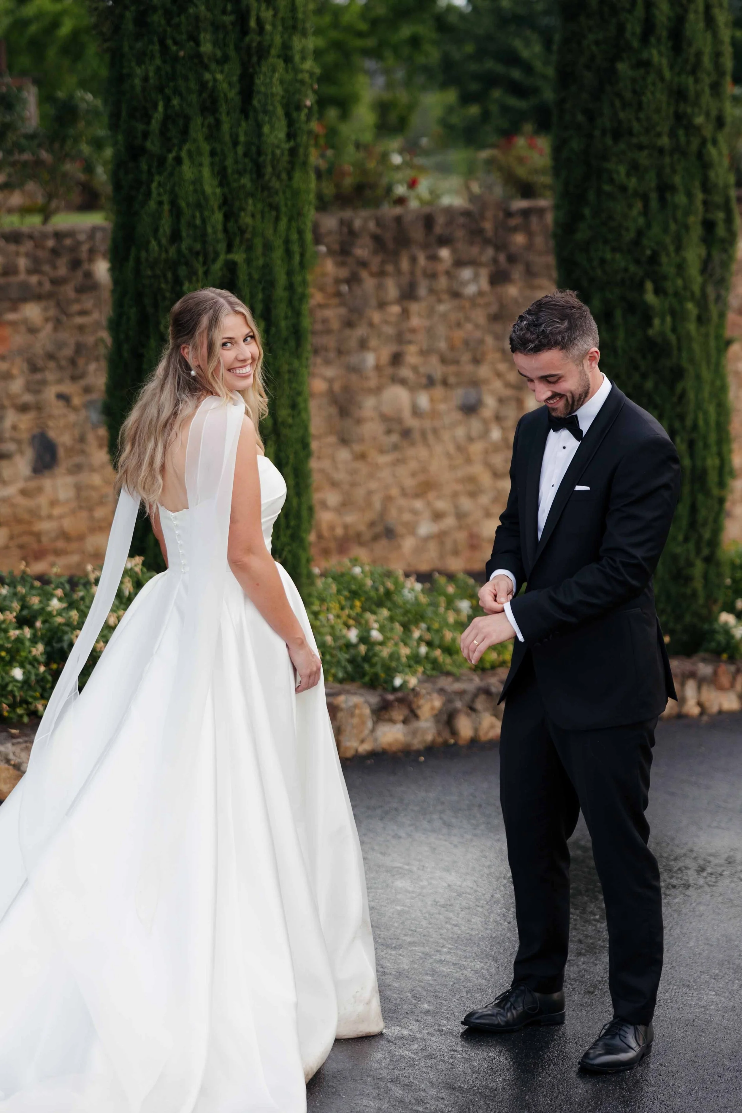 A bride in a white wedding gown and a groom in a black tuxedo smile, standing outdoors near a stone wall with tall green cypress trees in the background.