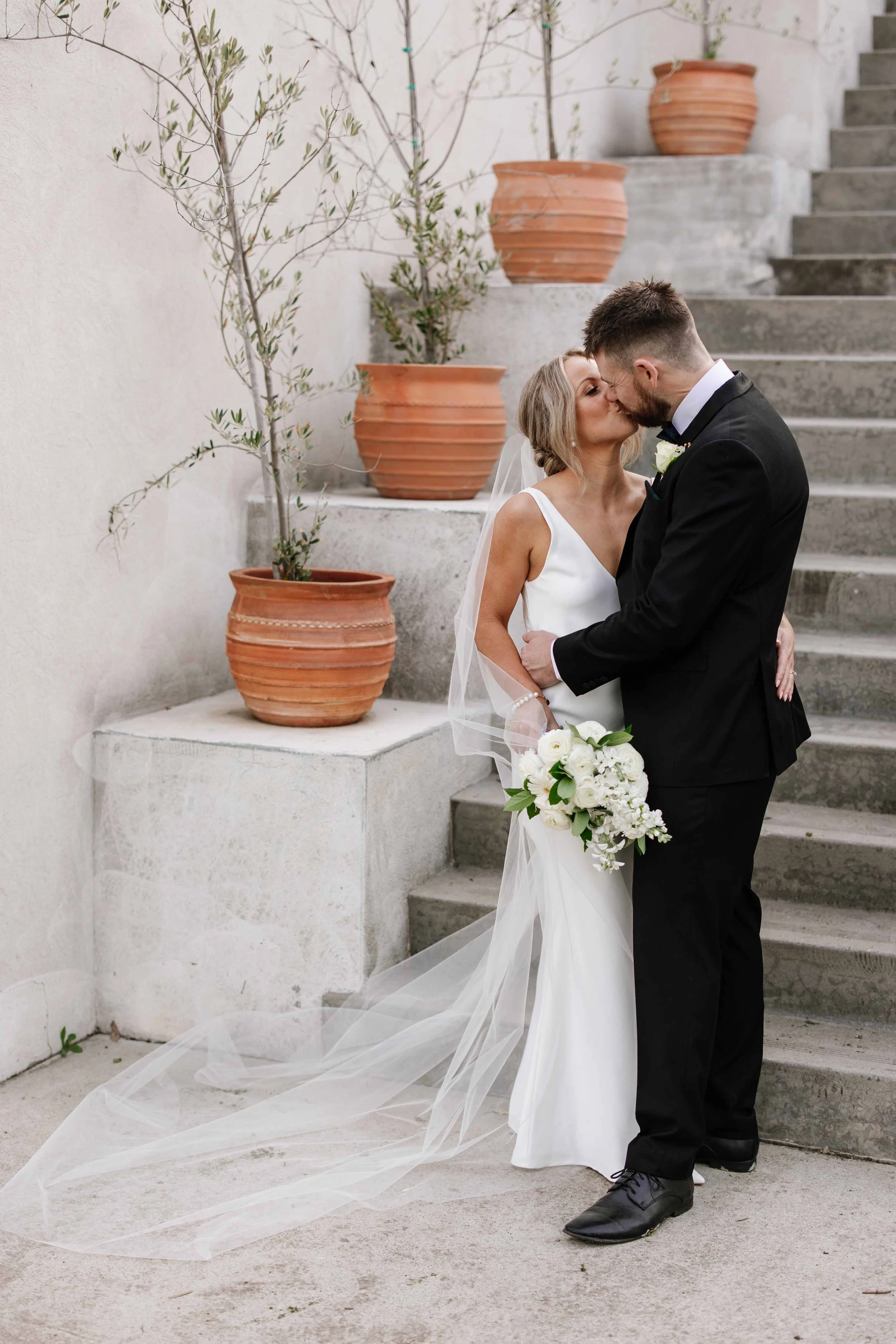 A newlywed couple sharing a kiss outdoors, with the bride in a white wedding dress holding a bouquet of white flowers, and the groom in a black tuxedo, standing on concrete stairs next to potted plants.