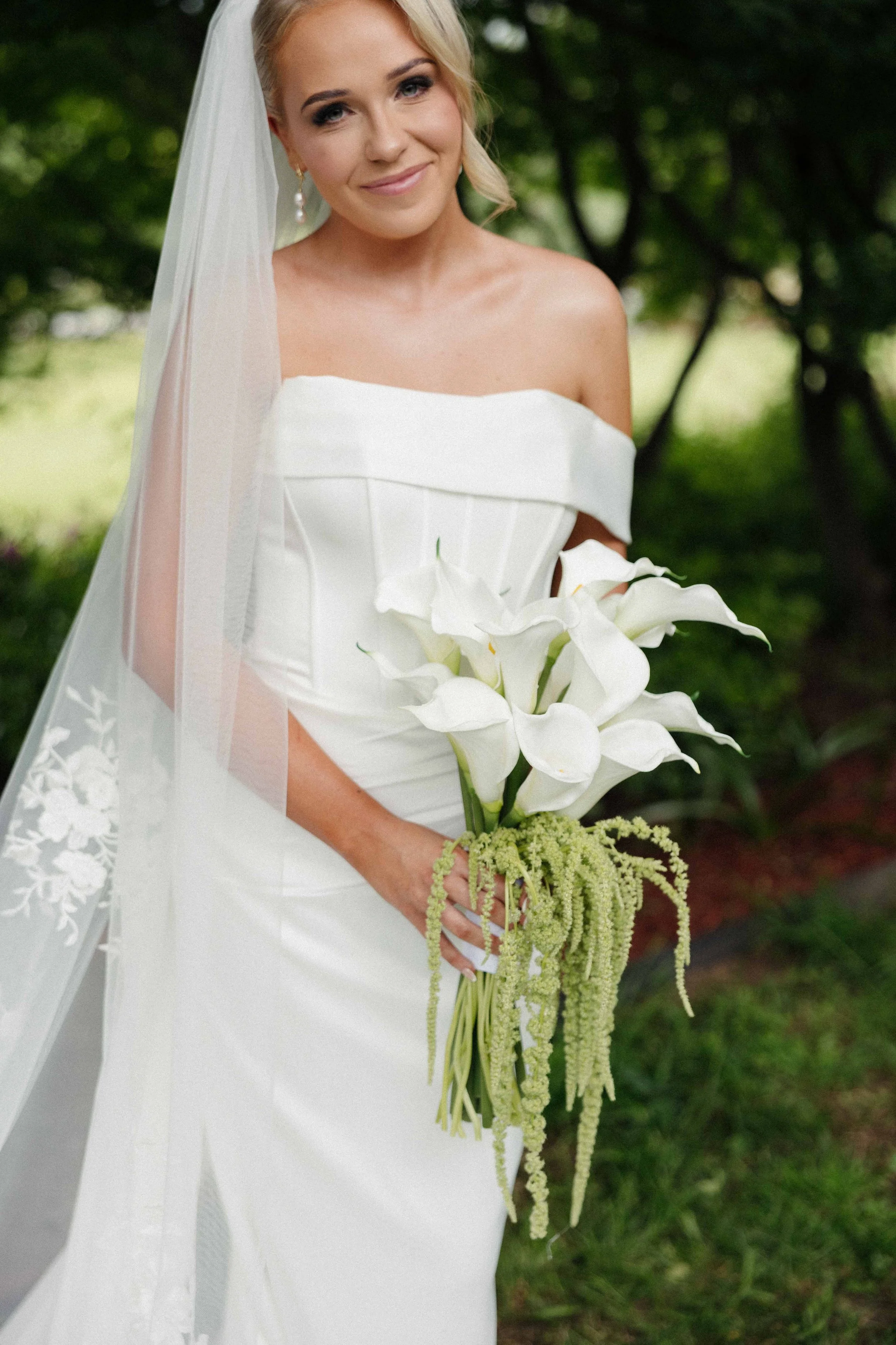 A bride in a white wedding dress holding a bouquet of white calla lilies with greenery, outdoors near trees.