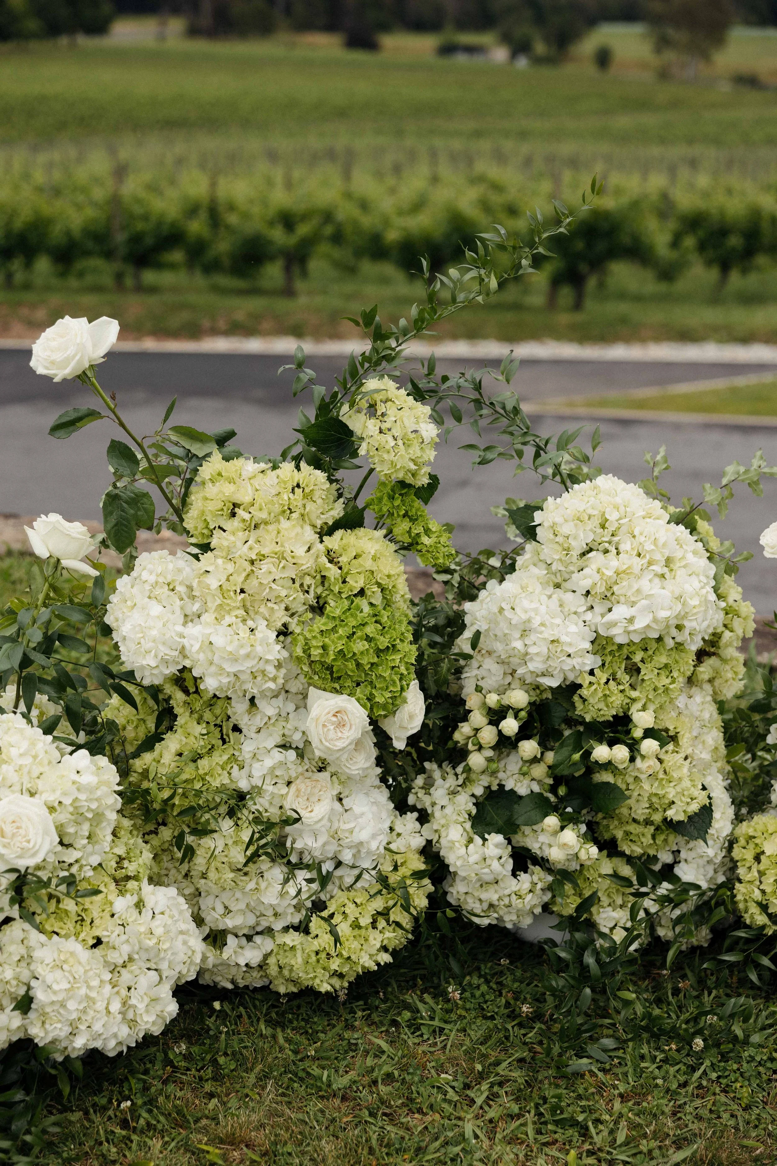 A large arrangement of white and light green hydrangeas and roses with green foliage, placed on grass near a paved area in a rural landscape with trees and fields in the background.