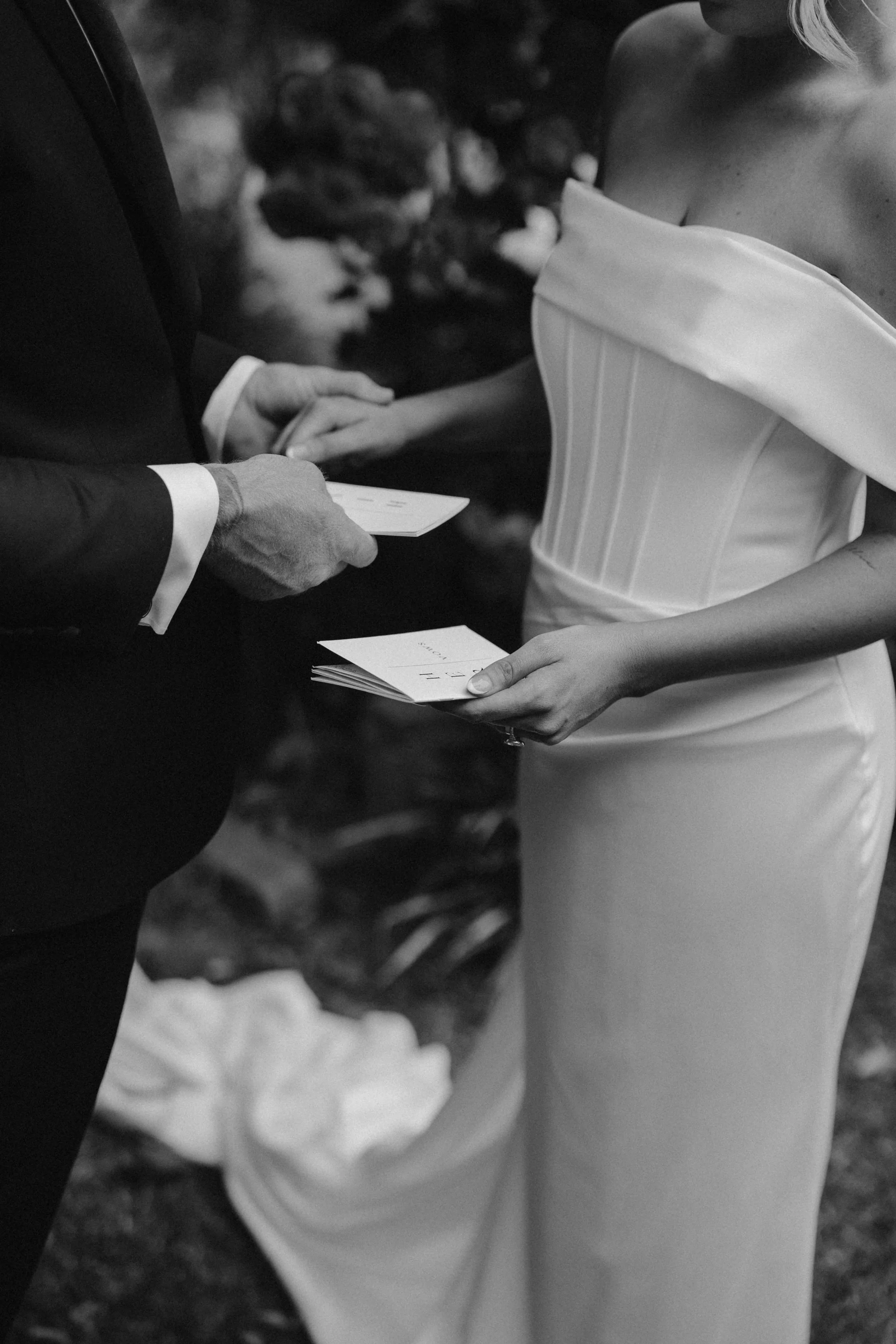 A black and white photograph of a wedding ceremony showing a close-up of a bride and groom exchanging vows and holding hands, with the groom holding a piece of paper and the bride holding some cards.