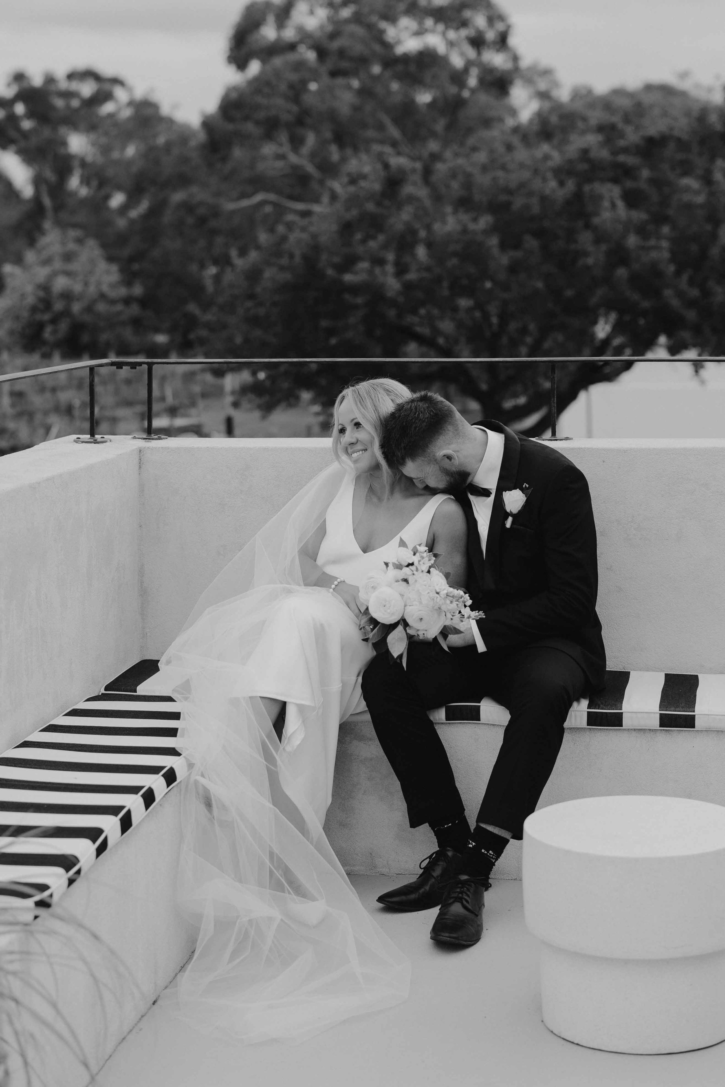 Black and white photo of a bride and groom sitting on a bench, with the groom whispering into the bride's ear, holding a bouquet of flowers, outdoors with trees in the background.