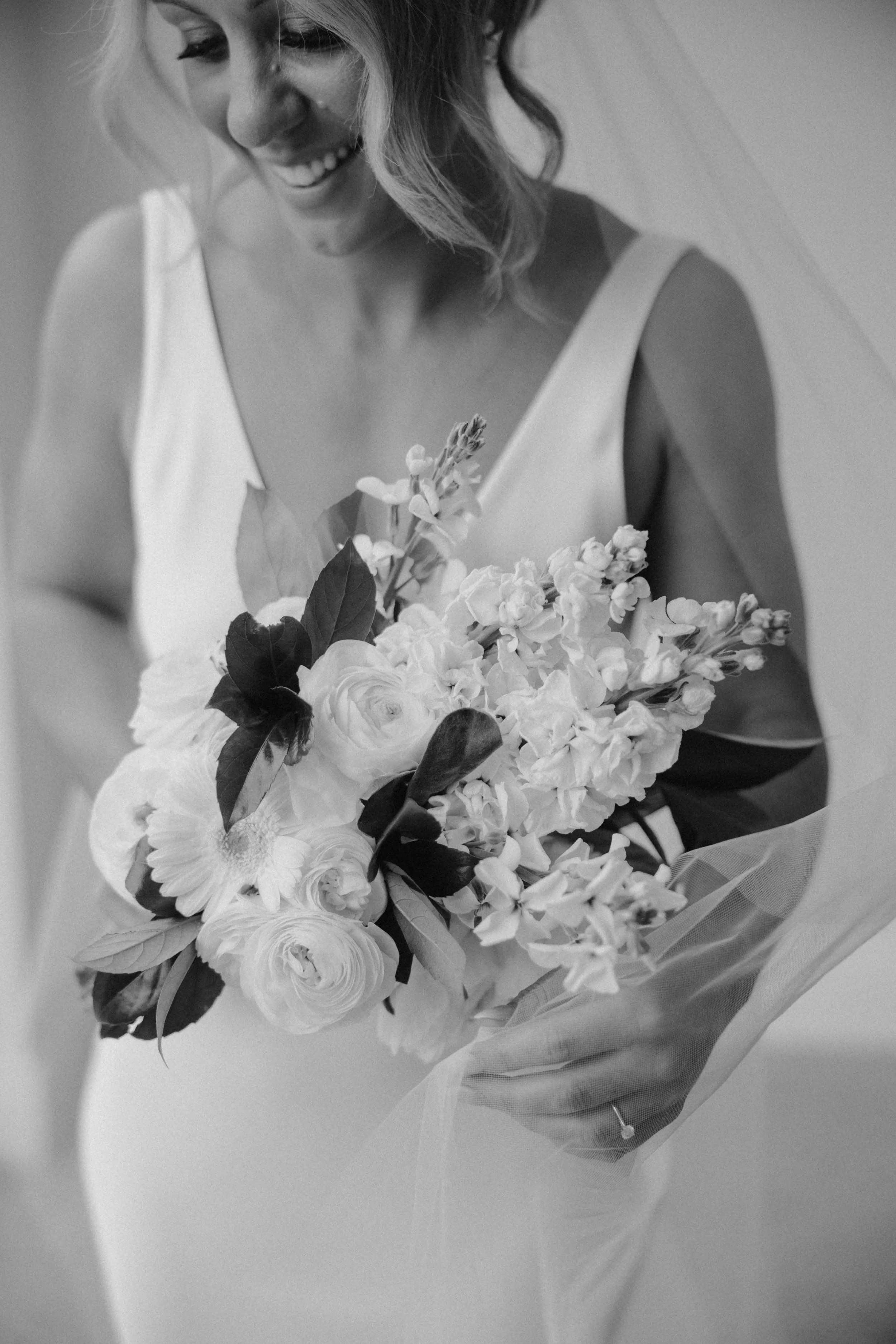 A smiling woman in a sleeveless dress holding a bouquet of flowers wrapped in sheer fabric.