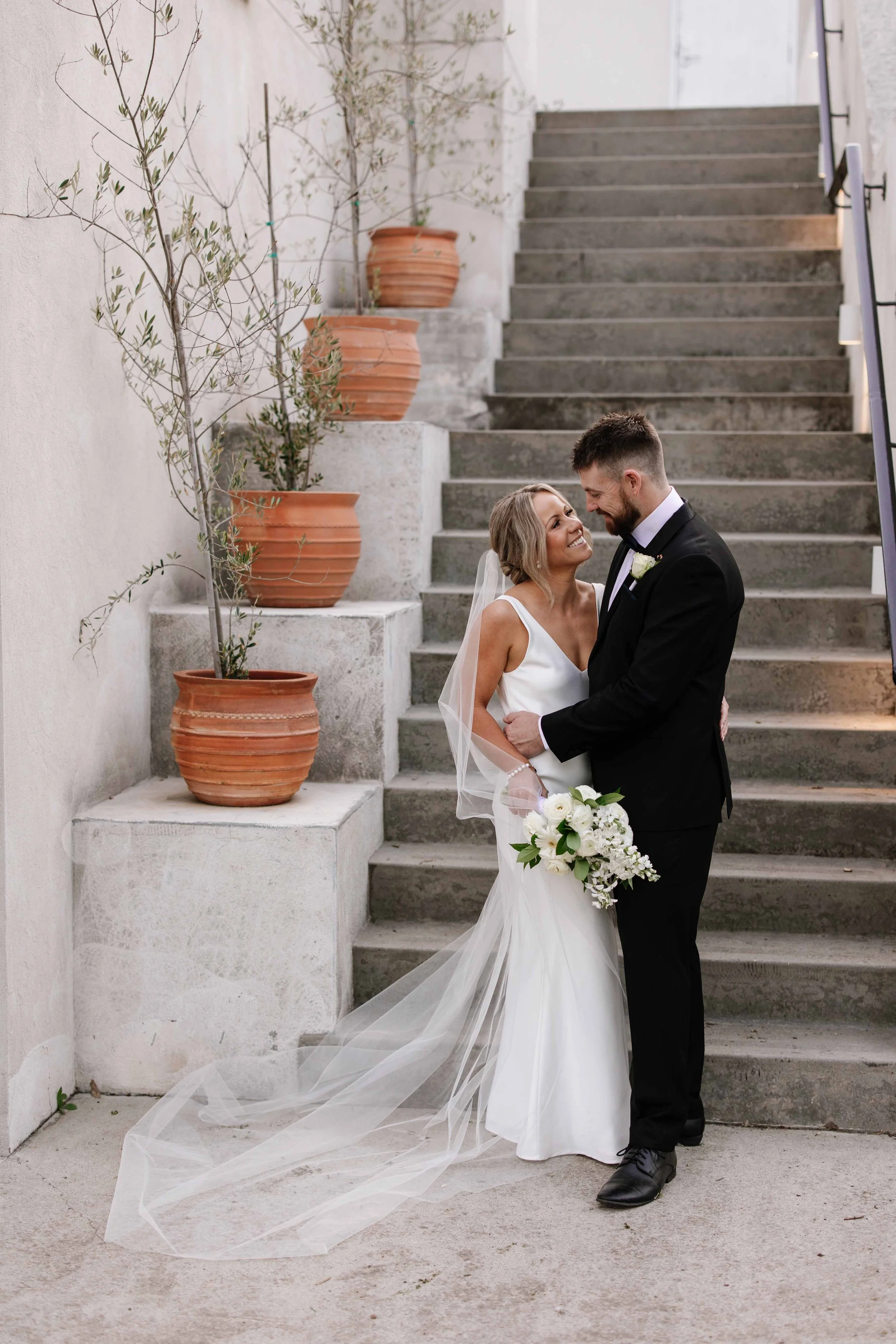 A bride and groom on a staircase, smiling at each other, with potted plants along the wall.