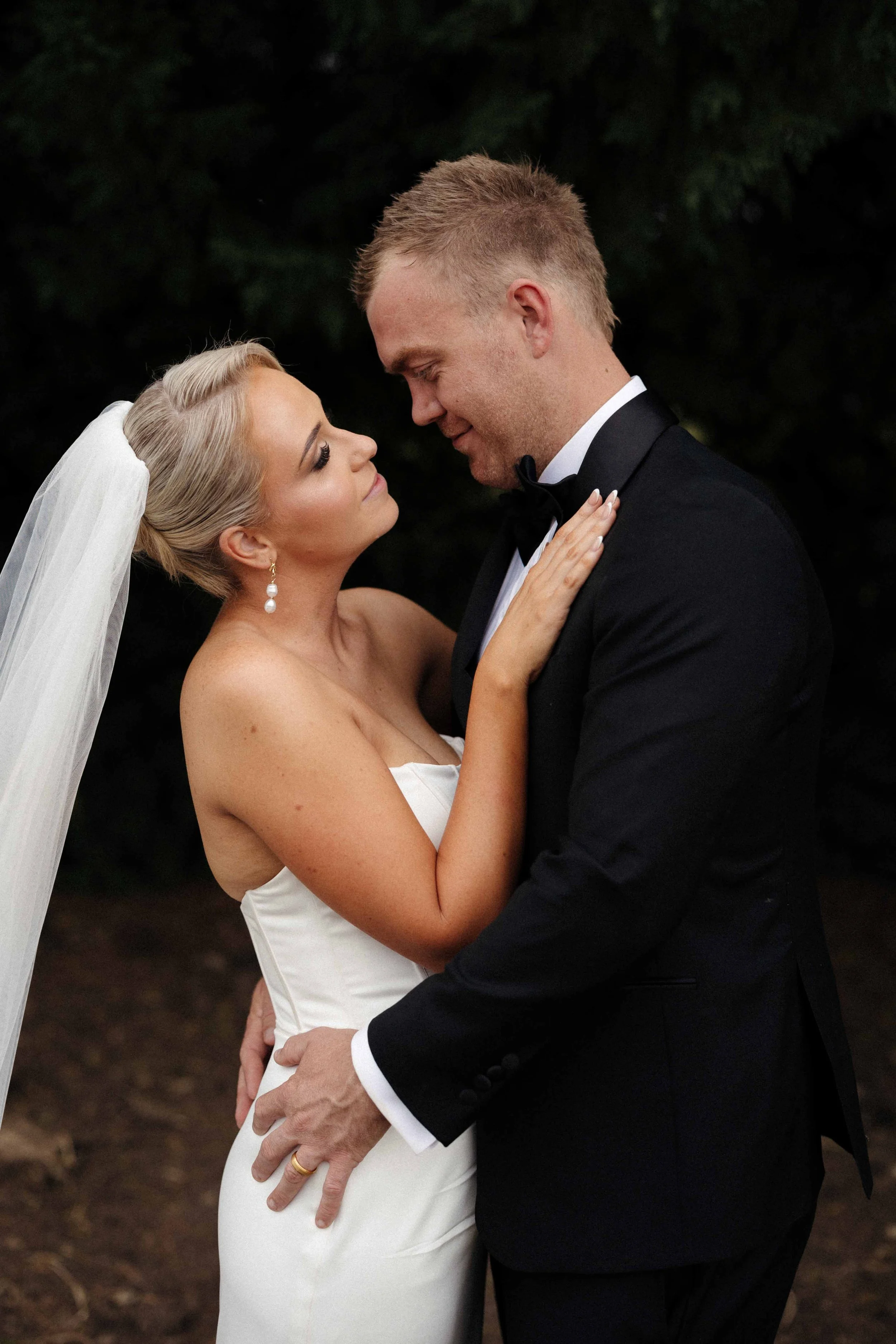 Bride and groom embracing outdoors, bride in white wedding dress and veil, groom in black tuxedo, dark trees in background.