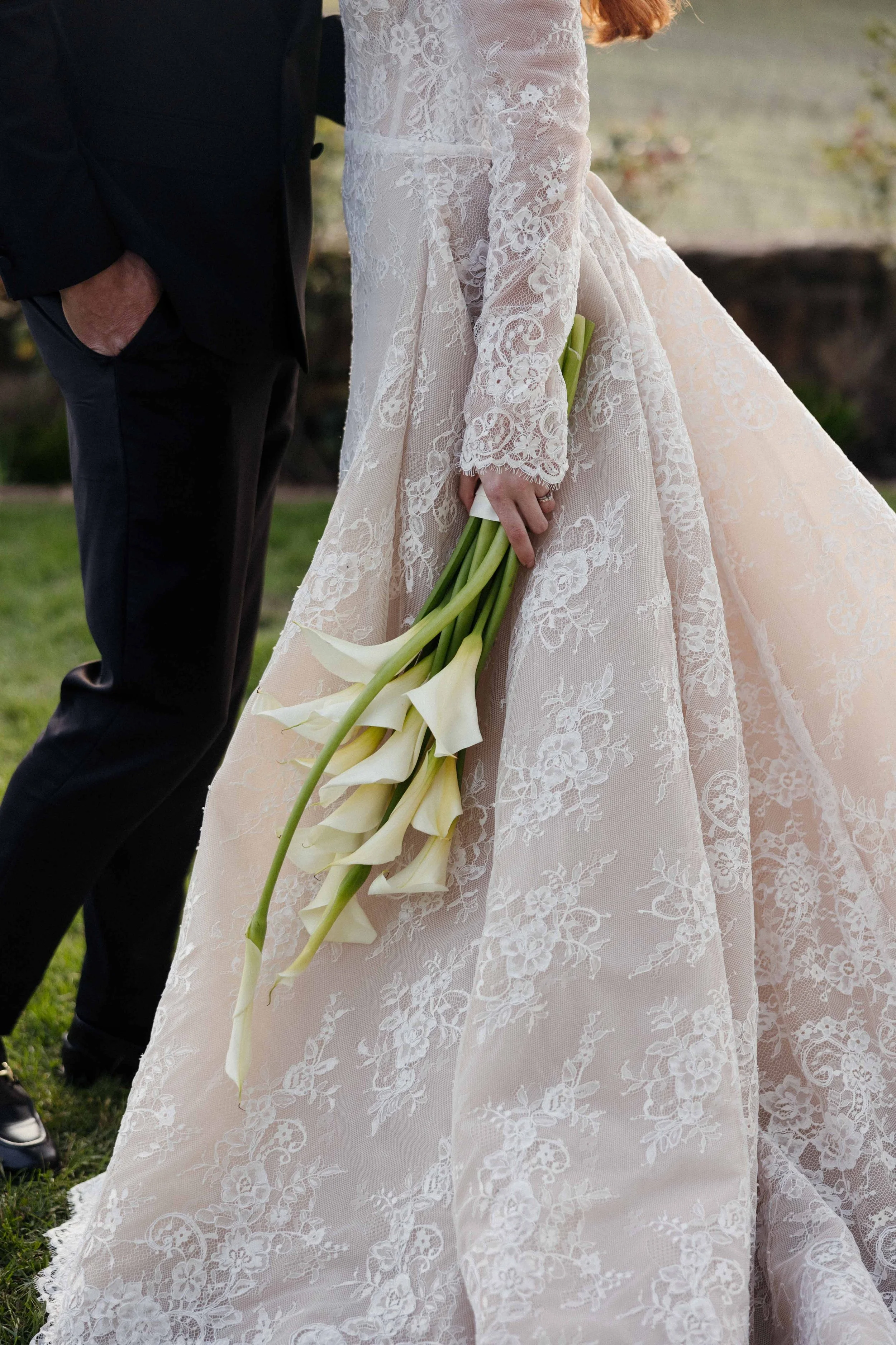 A woman in a lace wedding dress holding white calla lilies, standing next to a man dressed in black, outdoors on grass.