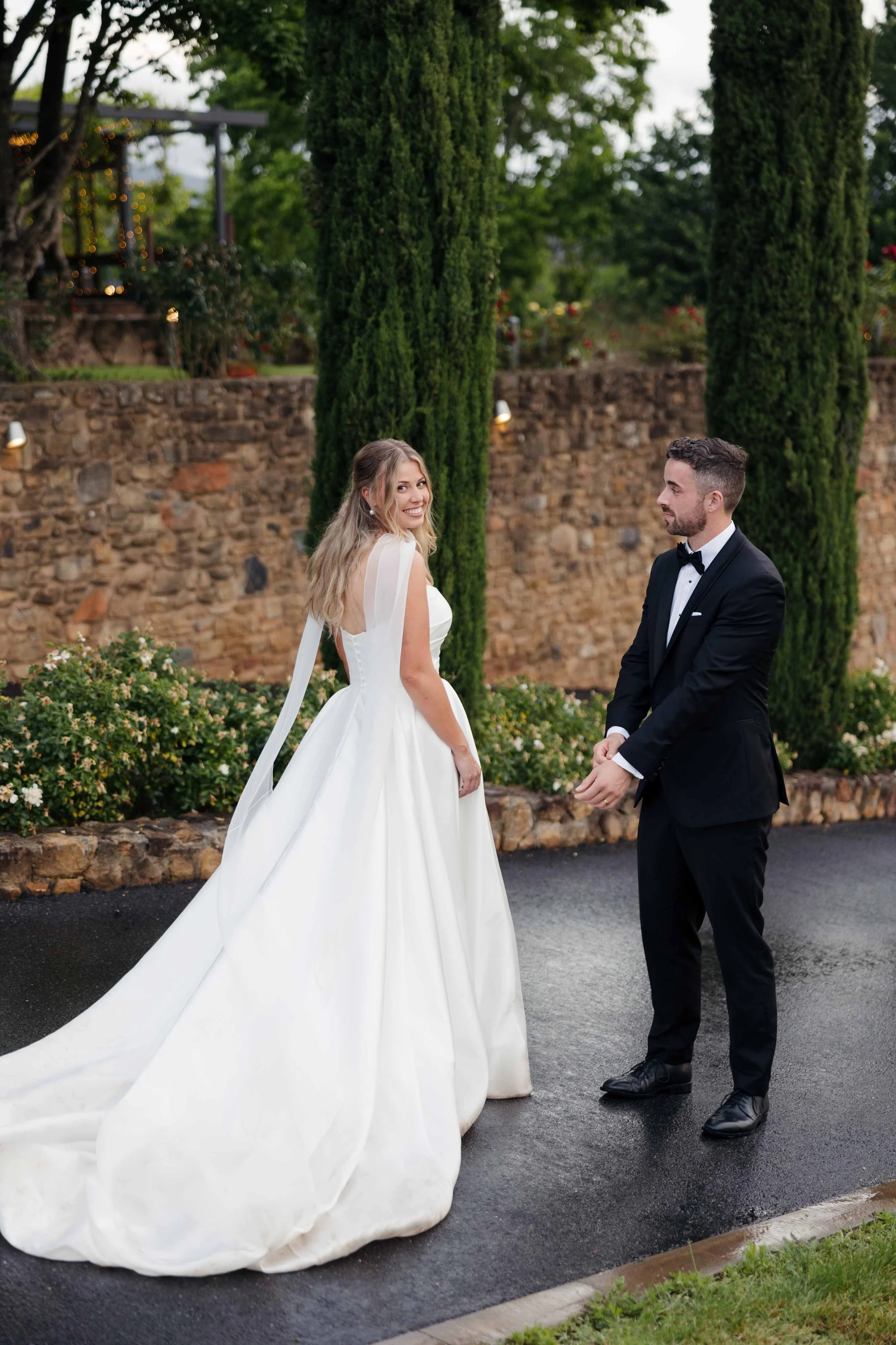 Bride and groom standing outdoors, couple on wedding day, woman in a white wedding gown, man in a black tuxedo, greenery and stone wall in background.