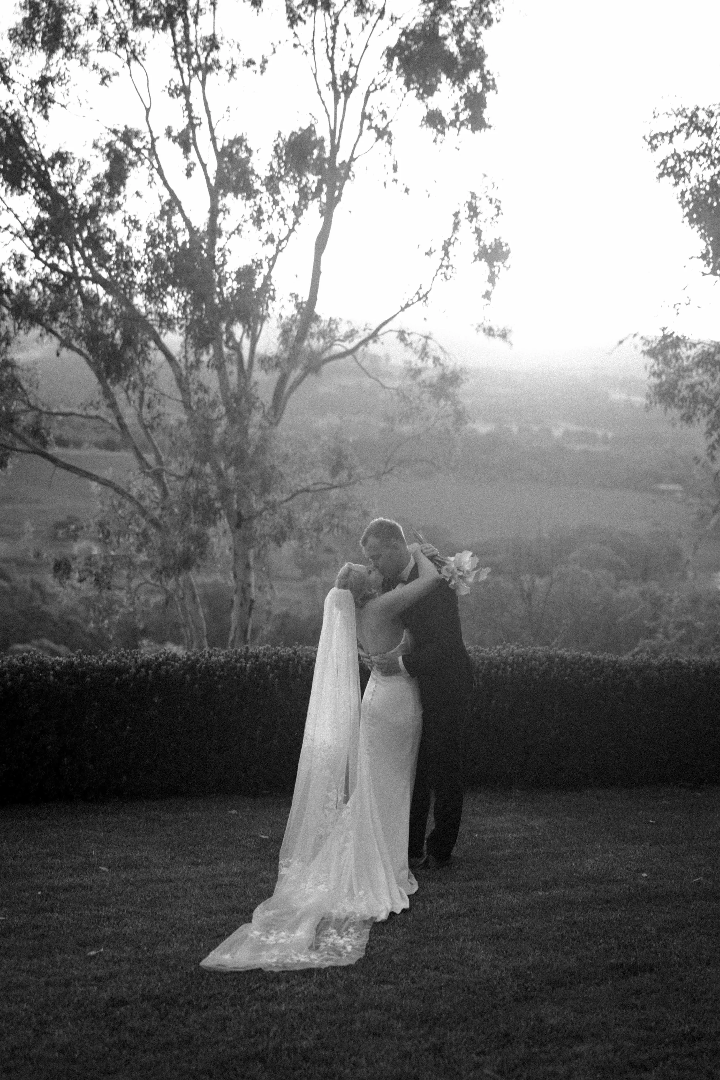 A black and white photo of a bride and groom embracing outdoors with trees and hills in the background.
