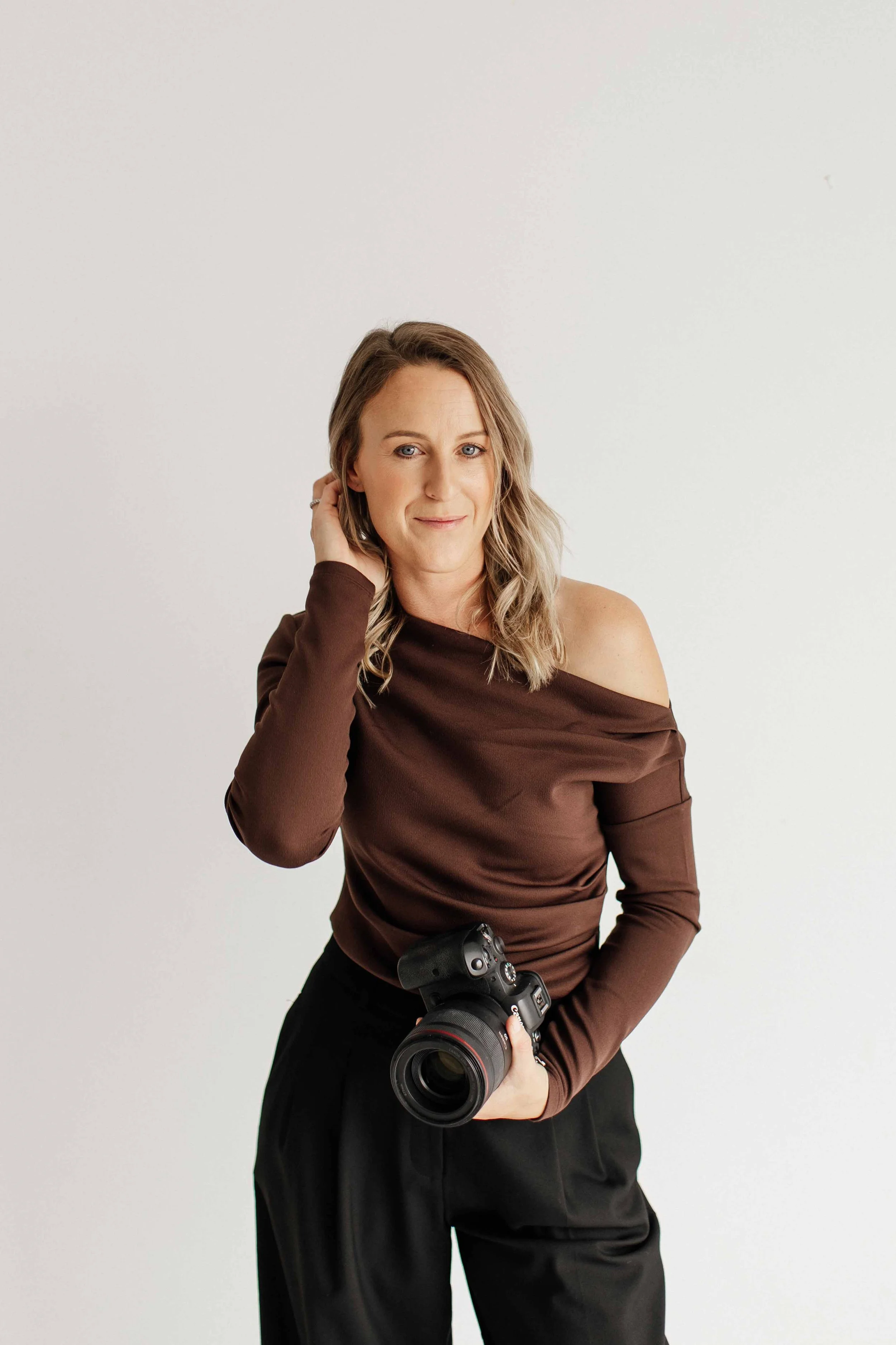 A woman holding a professional camera, wearing a brown off-shoulder top and black pants, standing against a plain white wall.