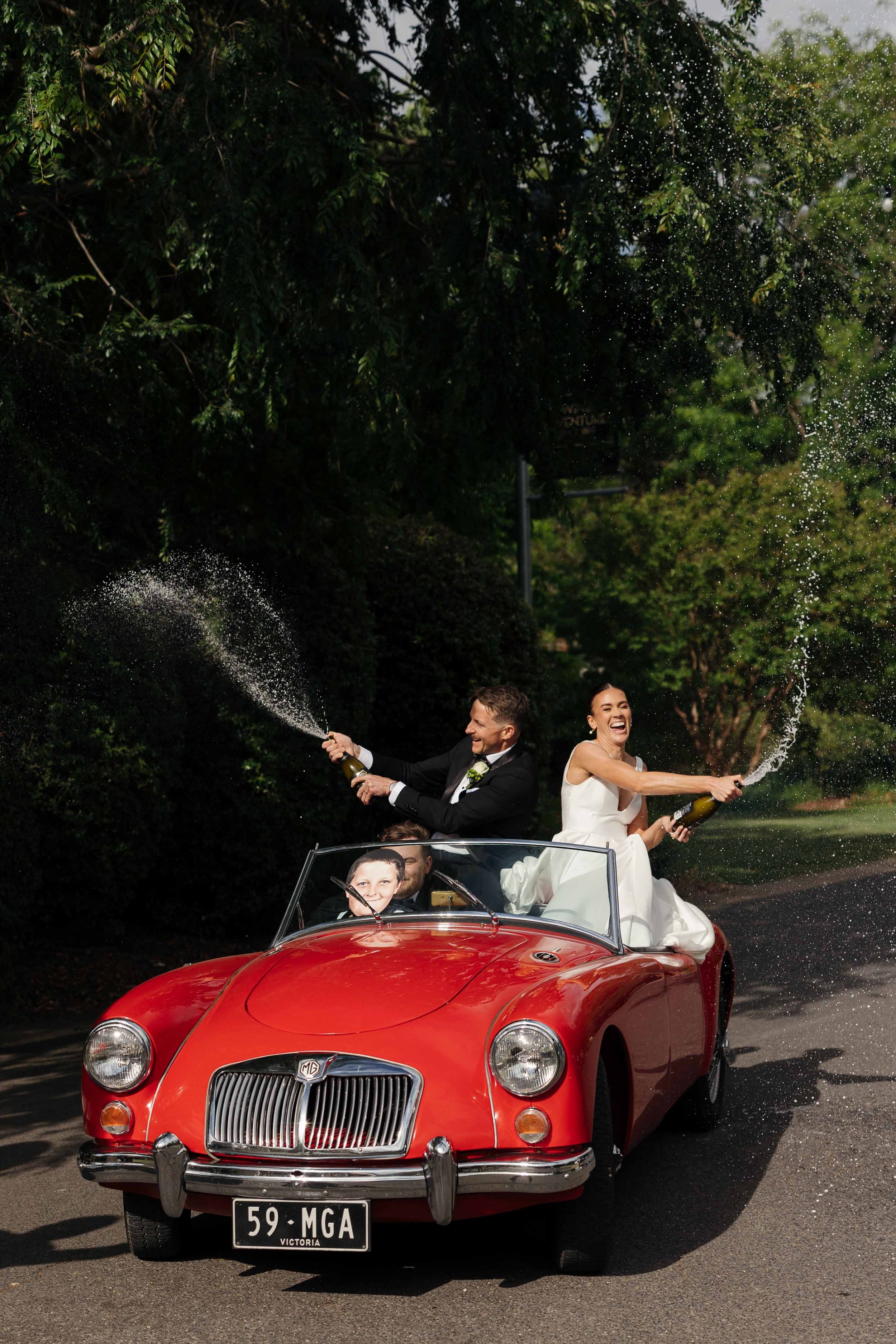 A newlywed couple celebrating in a vintage red MG convertible, spraying champagne as they drive down a tree-lined street.