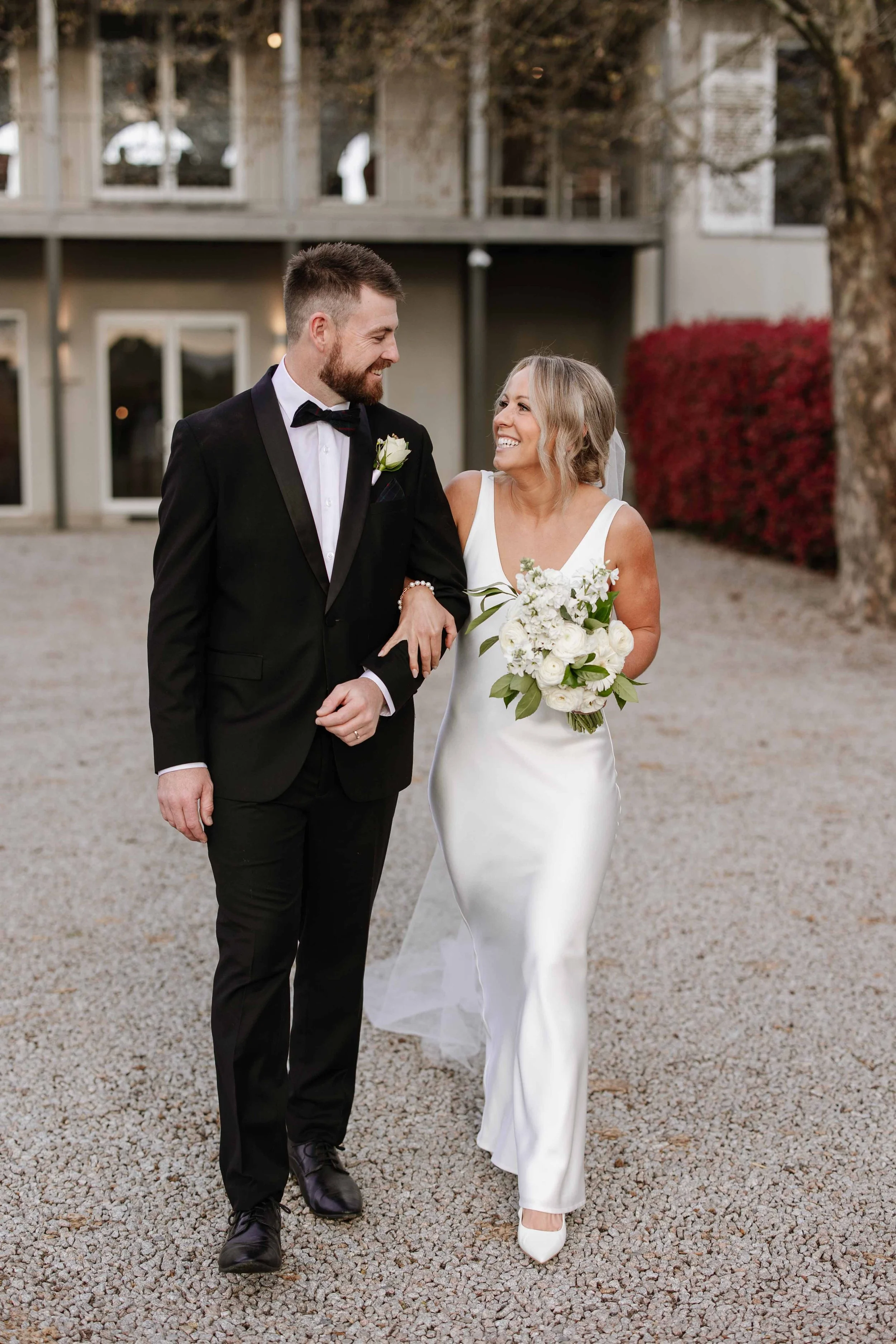 A bride and groom smiling and walking arm-in-arm outdoors during their wedding, with the bride holding a bouquet of white flowers and greenery.