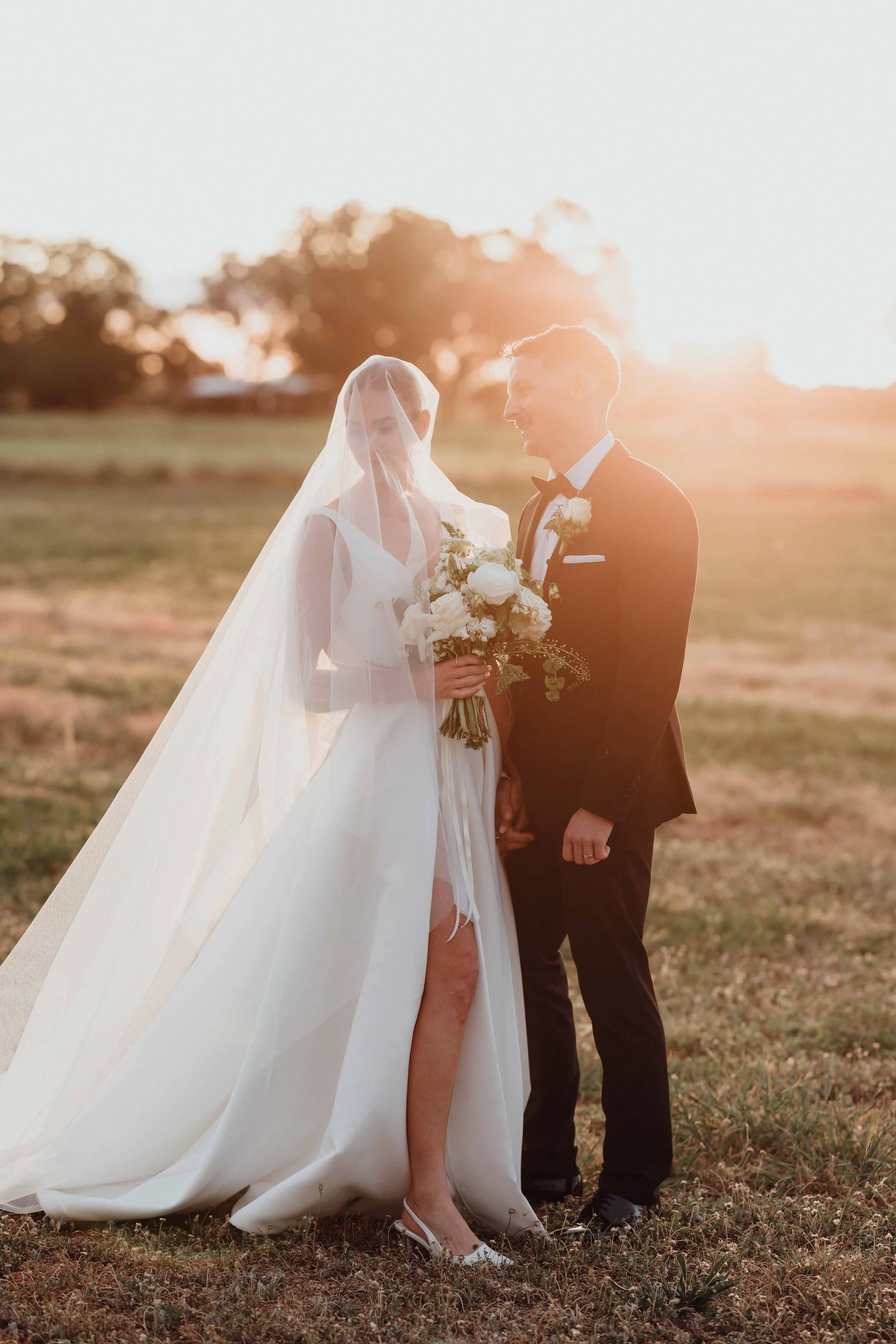 Bride and groom holding hands outdoors during sunset, dressed in wedding attire, with the bride wearing a long veil and white gown, and the groom in a black tuxedo.
