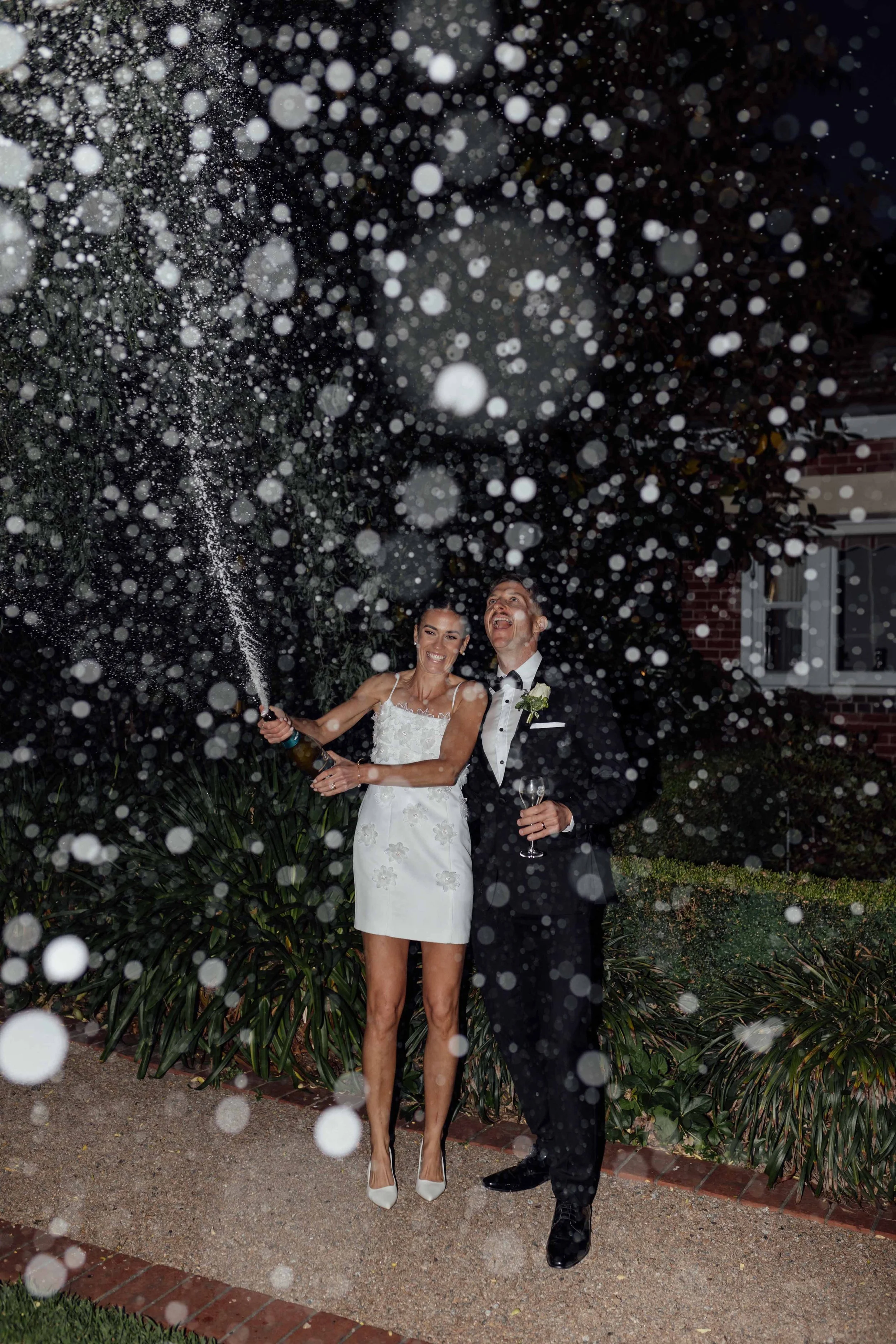 A newly married couple celebrating with champagne and bubbly at night outdoors, with champagne spraying from the bottle and confetti in the air.