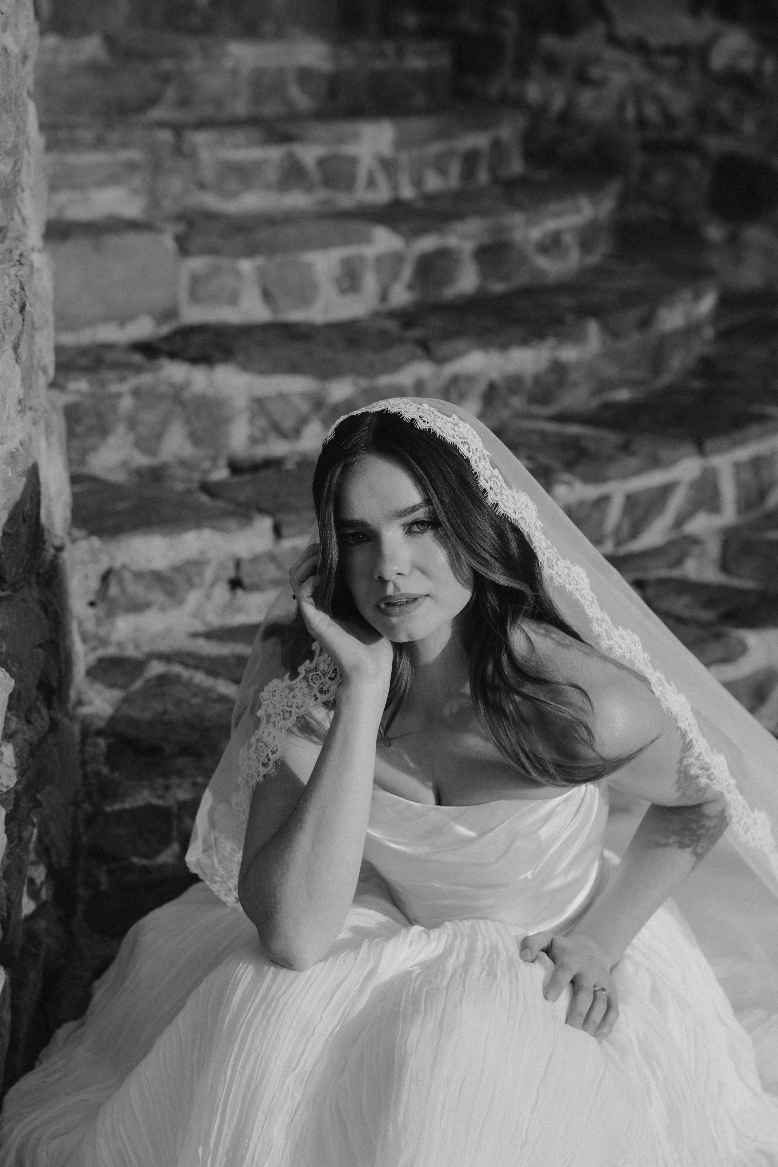 Black and white photo of a woman in a wedding dress with a lace veil, sitting on stone steps and resting her chin on her hand, looking at the camera.